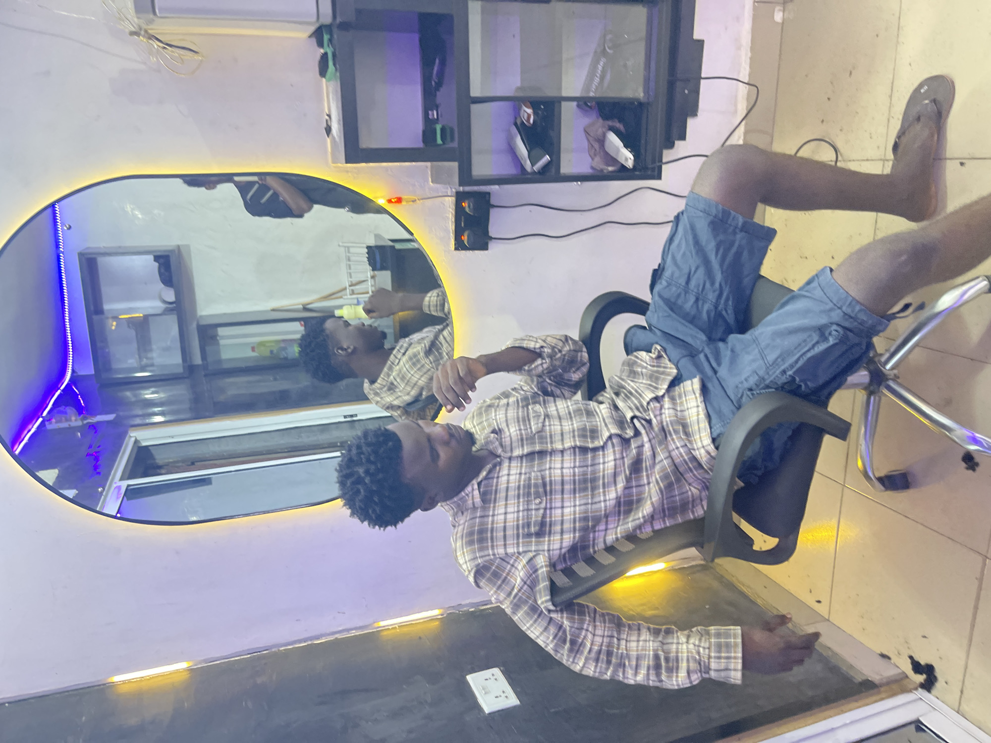 A young man with dark, curly hair is seated casually in a black and chrome swivel chair within an indoor space, likely a salon or barber shop in Unguwar Gabas, Nigeria. He is wearing a plaid long-sleeved shirt in shades of brown and white, and blue cargo shorts, with dark flip-flops on his feet. He looks downwards, appearing to be in a relaxed state or waiting.
Behind him is a large, oval mirror, dramatically lit with a warm yellow LED strip tracing its perimeter. To the left of the mirror, blue LED strip lighting adds to the ambient glow. The man's reflection is clearly visible in the mirror, showing him from a different angle, along with a container on a counter in front of him. Above the mirror, a dark wooden or metal shelving unit with multiple compartments holds various items, including what appears to be hair clippers with a green handle and white boxes.
The walls are light-colored, and the floor is tiled in a beige or light brown hue. Several electrical cables run along the walls and floor. An air conditioning unit is partially visible at the top of the frame. In the foreground, a white electrical outlet is visible on a dark surface. The lighting and setup suggest a modern grooming establishment. No clearly legible text is visible in the image.
