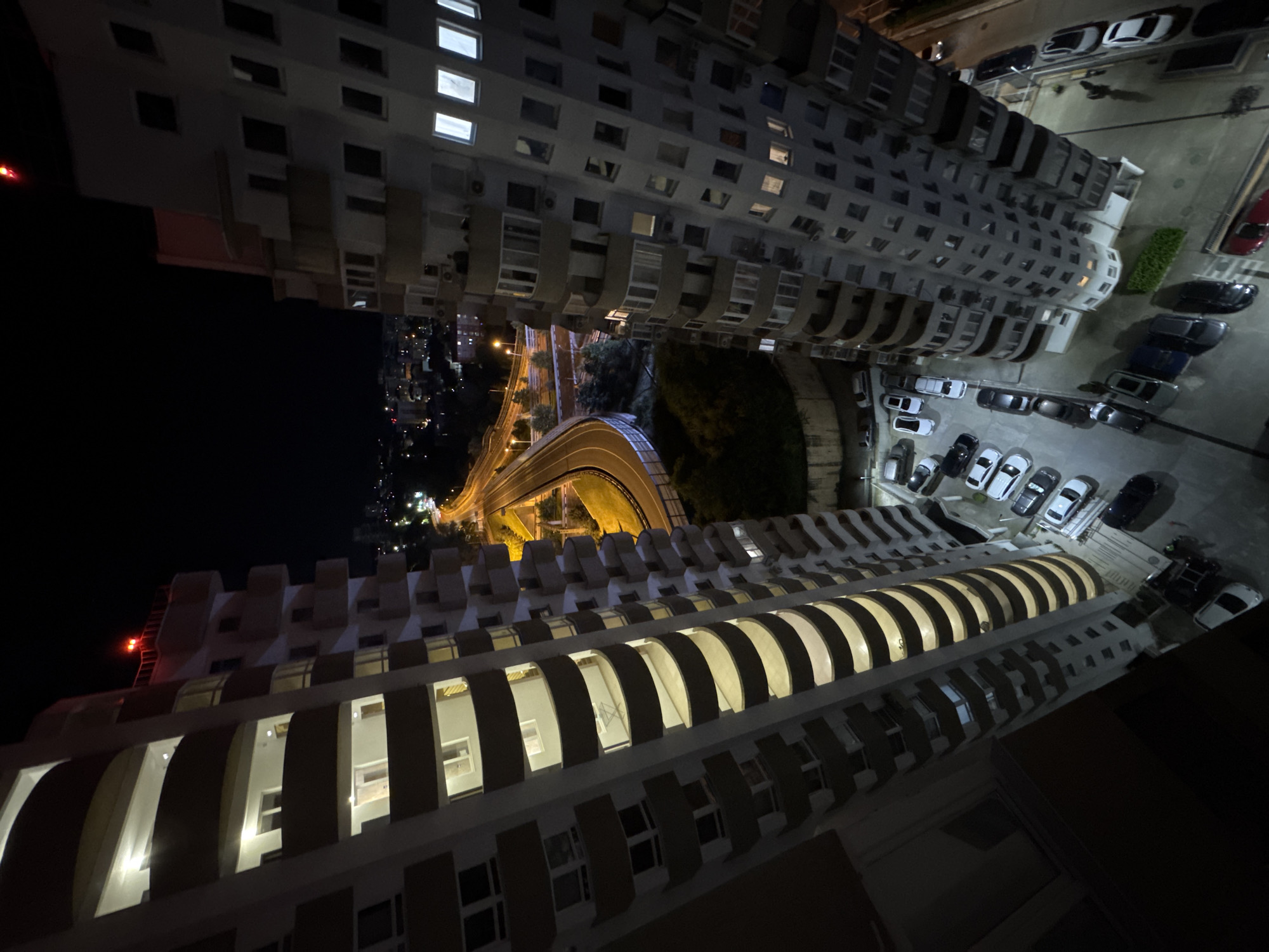 This image presents a detailed, high-angle nocturnal view of a modern urban environment in Sochi, Russia. The scene is dominated by two towering, multi-story residential buildings, likely apartment complexes, framing the composition.
The building on the left features a distinctive curvilinear facade with numerous balconies, many of which are illuminated, revealing interior spaces. Its structure is predominantly white with brown balcony railings. A set of red aviation obstruction lights is visible on its rooftop.
The building on the right is similar in height and architectural style, also exhibiting multiple illuminated windows and balconies. Numerous external air conditioning units are affixed to its facade. This building also displays red aviation obstruction lights on its rooftop.
Between and at the base of these buildings lies an asphalt parking lot, accommodating approximately two dozen parked passenger vehicles of various colors, including white, black, silver, and red. One individual is discernible standing near a parked car in the lower right quadrant of the parking area. Small rectangular beds of manicured greenery are interspersed within the parking lot.
Above the parking lot and between the buildings, a brightly illuminated, multi-lane highway or major road system curves from the left foreground towards the right background. The road is bordered by yellow guardrails and illuminated by numerous streetlights emitting a warm yellow-orange light. Below the elevated road section, additional green foliage and landscaping are visible.
In the distant background, a sprawling cityscape extends under a dark night sky, marked by countless scattered lights indicating a dense urban settlement. The overall scene is characterized by artificial lighting against the dark backdrop of night, creating a strong contrast and highlighting the urban infrastructure.