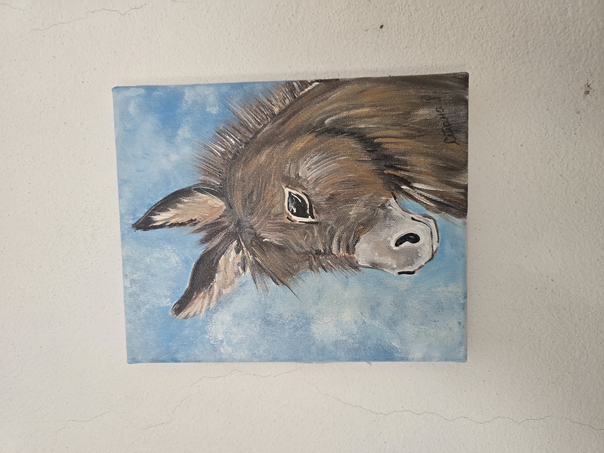 A square canvas painting of a donkey's head is hung on a textured white wall. The donkey is facing left, with its head tilted slightly down and its ears perked up. The painting is done in acrylics, with shades of brown, white, and black for the donkey's fur, and a light blue and white for the sky background. A signature in dark cursive is visible in the lower right corner of the painting. The wall has a rough, stucco-like texture and shows some faint cracks. The lighting appears to be overhead, casting a slight shadow on the wall to the left of the painting. There are no other objects or people visible in the image.