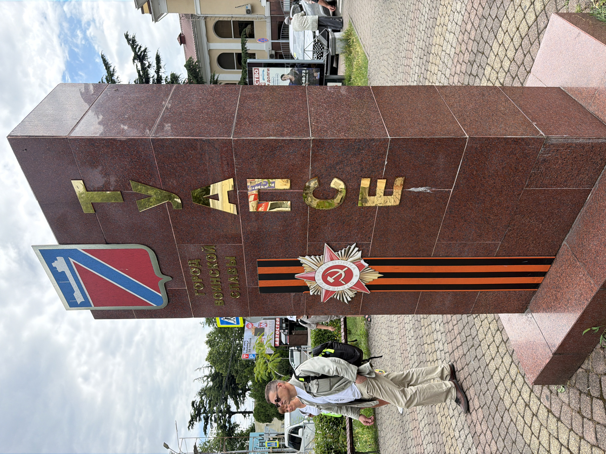 The image shows a monument in the city of Tuapse, Russia. The monument is made of red granite and has gold lettering on its side that spells out "ТУАПСЕ". At the base of the monument is a red and black ribbon, resembling the St. George's ribbon, and a red star with a hammer and sickle emblem, symbolizing Soviet military glory. Above the lettering, there is a coat of arms featuring a ship and a red and blue shield.
In the foreground, a man in a light-colored jacket and pants is walking past the monument. He is wearing sunglasses and carrying a backpack. Behind him, a woman in a white top and shorts is also walking. In the background, there are trees, buildings, and other people. A street sign is visible in the background, partially obscured by the monument. A white van is parked on the left side of the image. The ground is paved with cobblestones. The sky is cloudy.