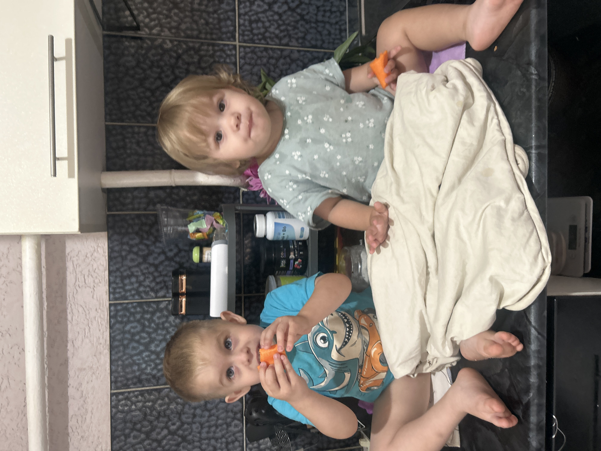 Two young children are sitting on a black countertop in a kitchen. The child on the left, a boy with blond hair, is wearing a blue t-shirt with a shark graphic and is holding an orange piece of food to his mouth. The child on the right, a girl with blond hair, is wearing a light blue floral-patterned shirt and is holding a similar orange piece of food. A beige blanket is draped over their laps. In the background, there are kitchen cabinets, a tiled backsplash, and various containers and bottles on shelves. The lighting suggests an indoor setting.