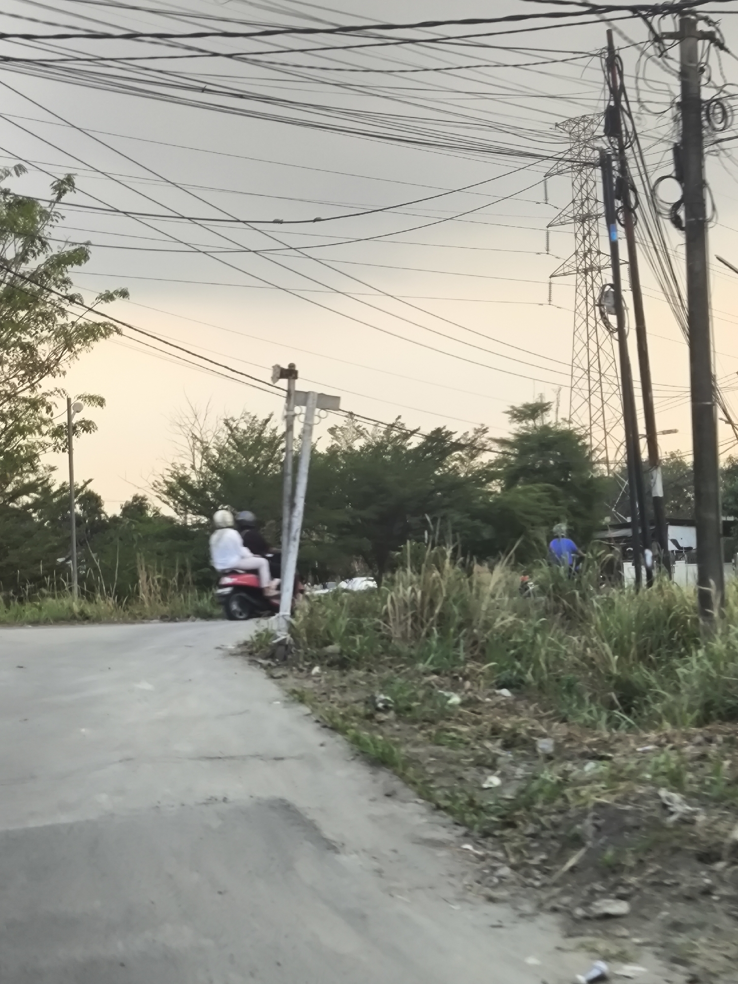 The image depicts a scene in Cilegon, Indonesia, characterized by a power line infrastructure and a paved road. In the foreground, a concrete road curves to the left. On the side of the road, tall grasses and some debris are visible. Further along the road, two individuals are riding a red scooter; the passenger is wearing a white helmet and a light-colored top, while the driver is wearing a dark helmet and top. To the right of the road, a wooden pole with electrical wires and insulators stands prominently. In the background, a large, lattice-like electrical tower can be seen, along with more electrical poles and a dense growth of trees. A person in a blue shirt is visible further down the road, near the electrical tower. The sky is overcast with a pale, yellowish hue, suggesting it might be early morning or late afternoon. The overall impression is one of an urban or semi-urban periphery with functional, yet somewhat cluttered, utility infrastructure amidst natural vegetation.