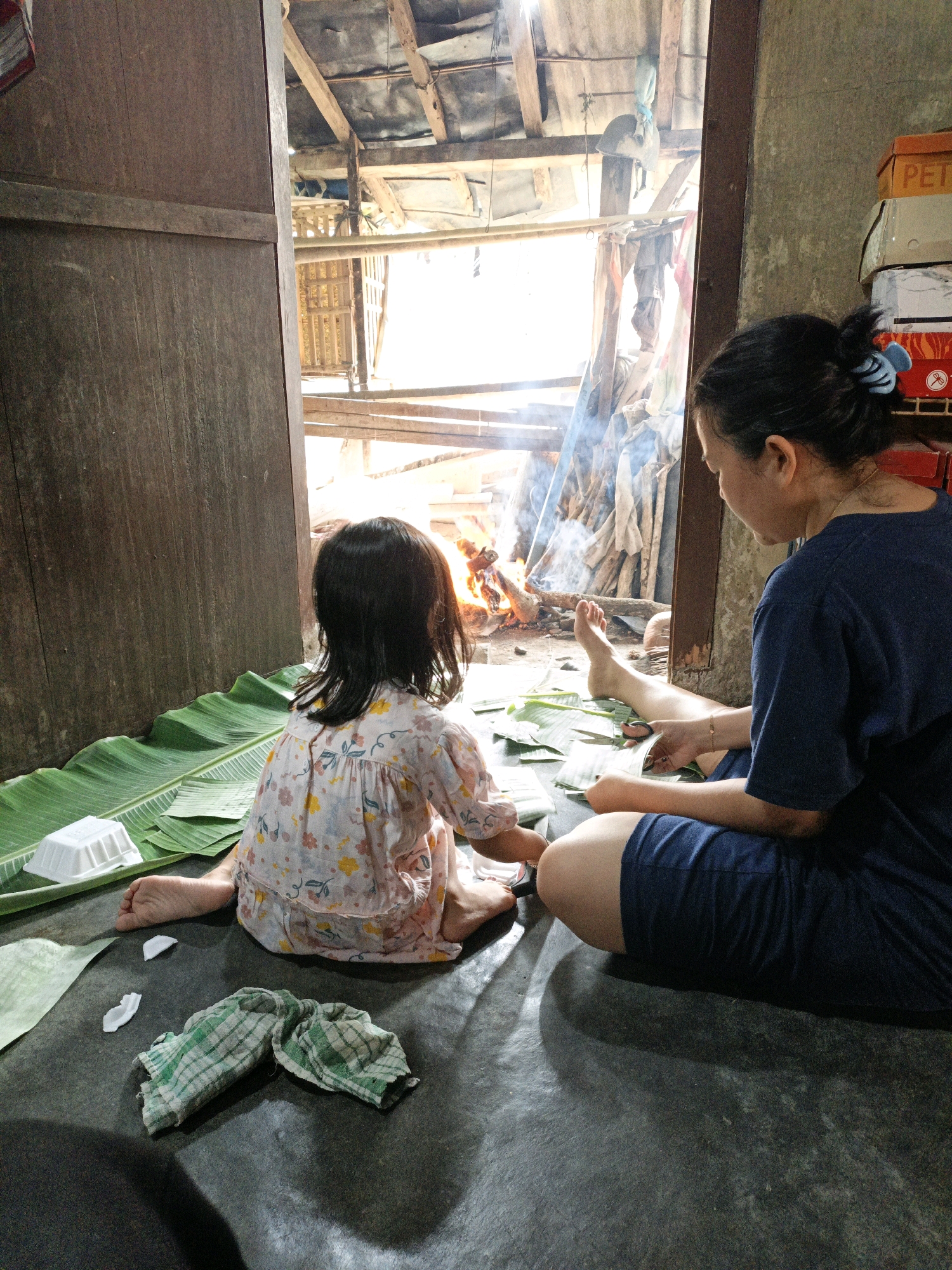 In Bunut, Indonesia, a woman and a young child sit on a dark floor, surrounded by large green banana leaves. The woman, wearing a navy blue shirt and shorts, is actively cutting the leaves with scissors. The child, in a floral dress, faces away from the camera, looking towards a fire burning in the background. The environment appears to be an open-air structure with a wooden ceiling and corrugated metal roofing, suggesting a rural or less developed setting. Smoke from the fire drifts upwards, partially obscuring the view of stacked materials and bamboo structures outside. The scene is lit by natural light streaming in from the opening. A white food container and a checkered cloth are also visible on the floor.