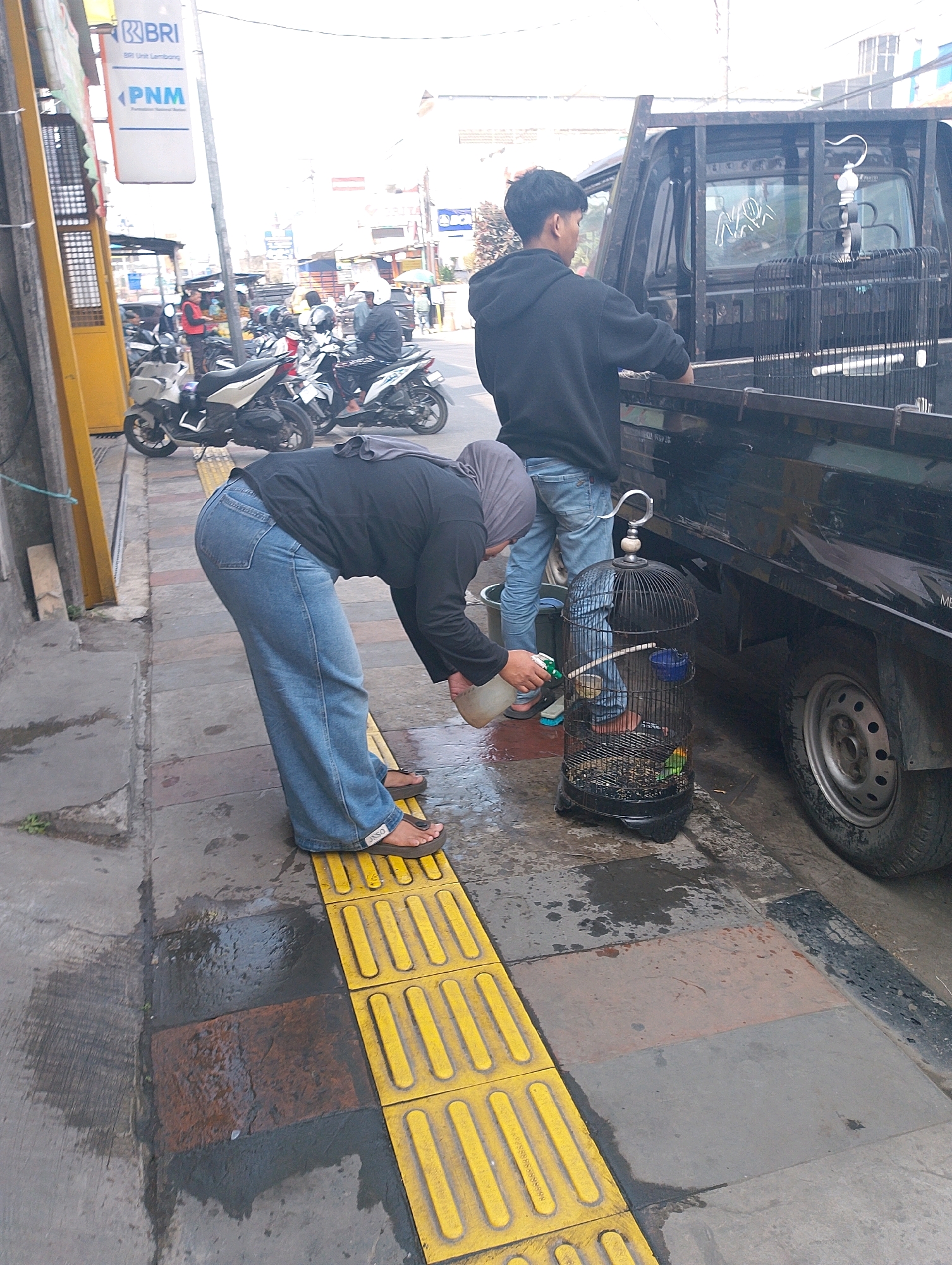 In Lembang, Indonesia, a person wearing a grey hijab and loose blue jeans is bent over, spraying a green liquid from a white plastic bottle into a black birdcage. The birdcage contains at least one small green and yellow bird. To the right of the birdcage, the tire and wheel well of a black pickup truck are visible. Behind the birdcage, a young man in a black hoodie and blue jeans stands near the truck. The scene is on a wet, paved sidewalk next to a street with parked motorcycles. Yellow tactile paving is present along the edge of the sidewalk. A sign for BRI (Bank Rakyat Indonesia) and PNM (Permodalan Nasional Madani) is visible in the background, suggesting a commercial area.