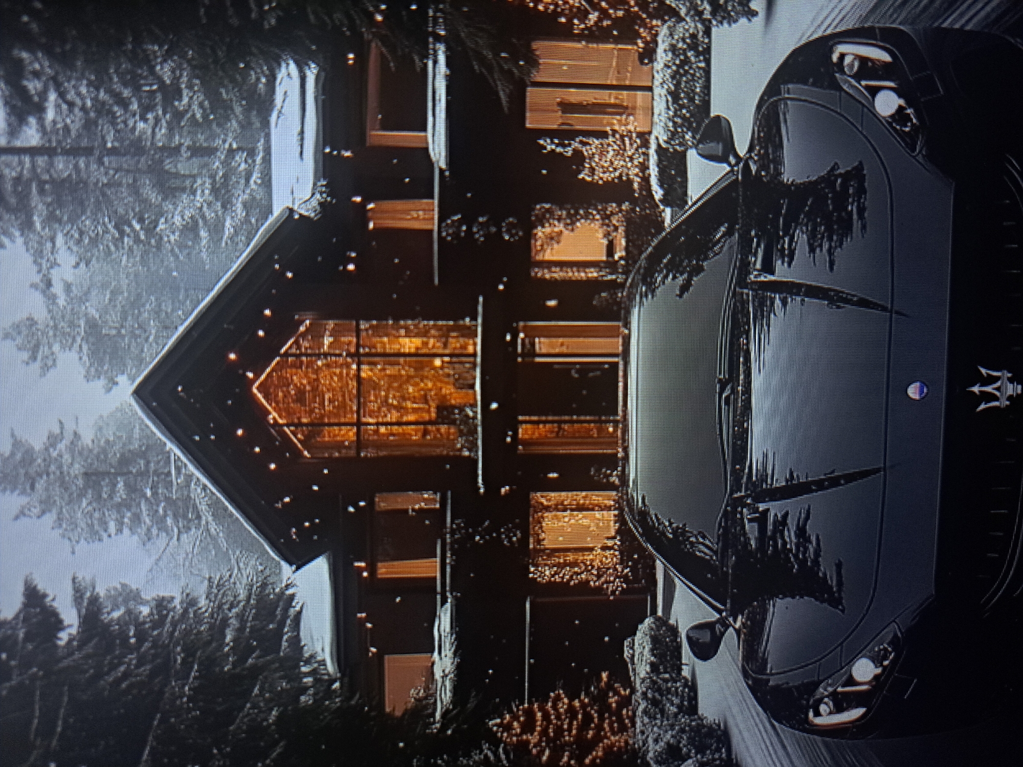 The image depicts a modern, dark-colored house with a large A-frame window facing the viewer. The interior lights illuminate the windows with a warm, orange glow. The house is surrounded by snow-covered trees and shrubs, suggesting a winter setting. A dark-colored Maserati automobile is parked in front of the house. It is unknown if the image is located in Yerevan, Armenia.