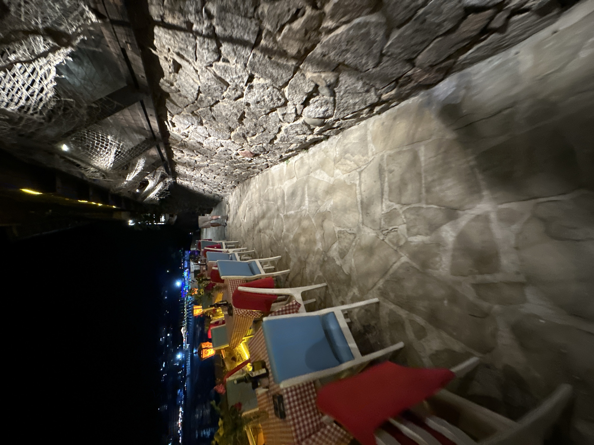 An outdoor restaurant setting at night is depicted. Tables are set with red and white checkered tablecloths and are adorned with red flowers and lanterns. Blue-backed chairs surround the tables. A stone wall runs along the right side of the frame, with a netting-like structure overhead. A pathway made of irregular stone slabs leads away from the dining area and alongside the wall. In the distance, lights from a city or town can be seen across the water. A person is walking away from the camera on the pathway. The overall atmosphere suggests a relaxed dining experience by the water.
