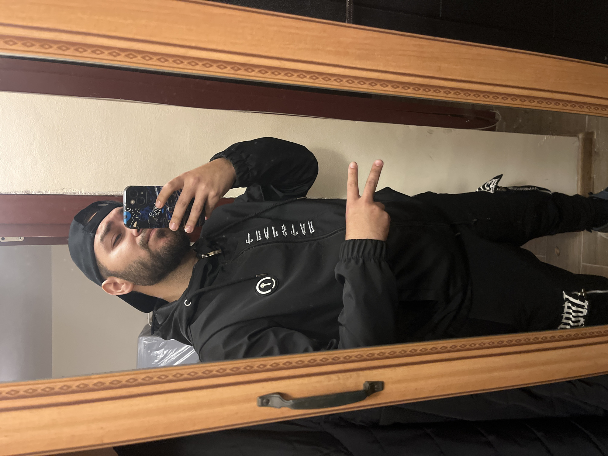 A man wearing a black baseball cap, black jacket, and black pants is taking a mirror selfie. He is holding a smartphone in his right hand, with the camera facing the mirror. He is making a peace sign with his left hand. The mirror has a light brown wooden frame with a decorative pattern. The background appears to be a room with light-colored walls. The location is not discernible from the image.