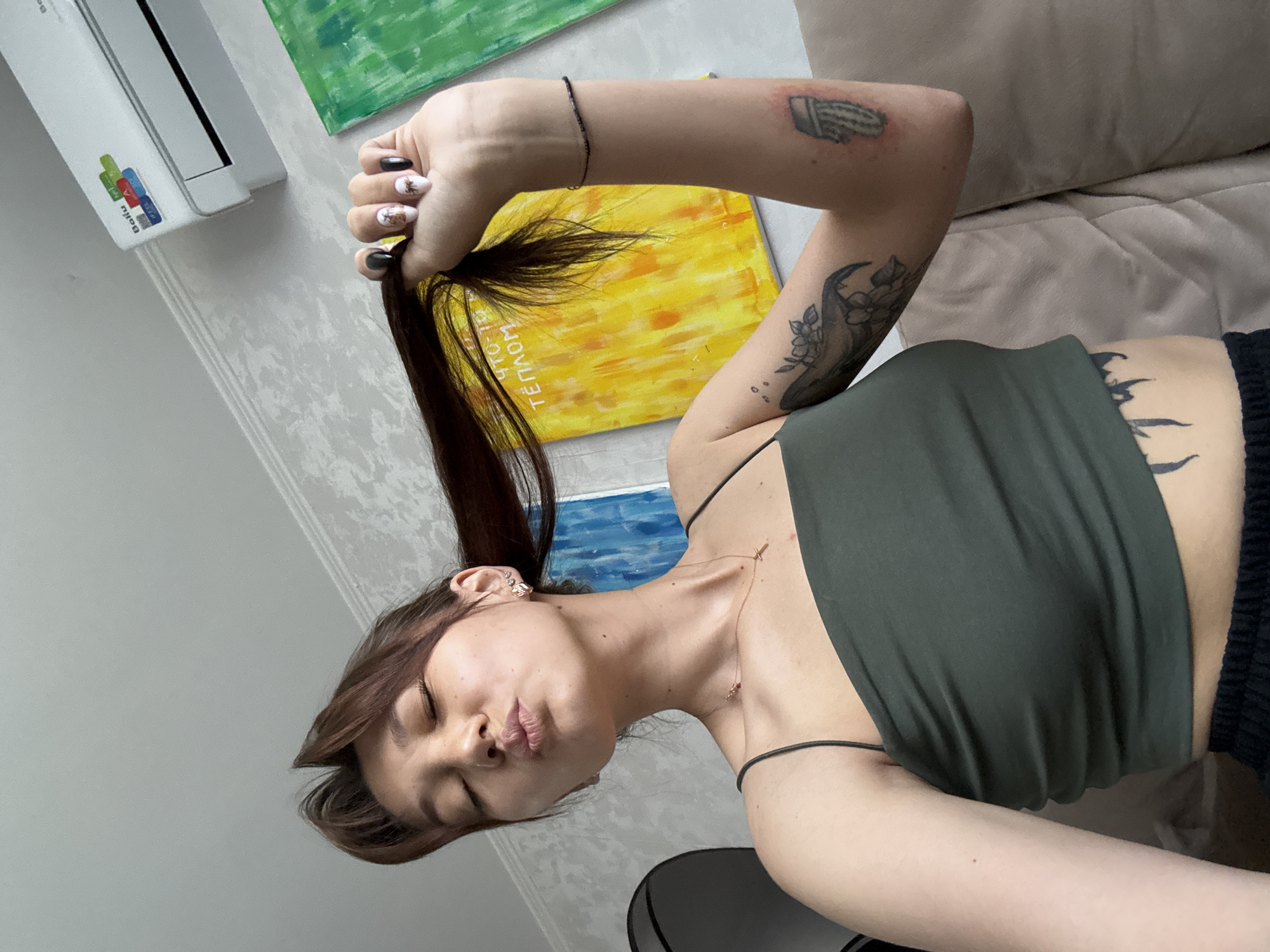 A young woman with brown hair pulled into a ponytail, with her eyes closed and lips pursed, is holding a lock of her hair in her right hand. She is wearing a dark green crop top and black pants. She has tattoos on her left arm and stomach. Behind her, there are two abstract paintings on the wall, one yellow and one blue and green. An air conditioner is mounted on the wall above the paintings. The scene appears to be an indoor setting, possibly a living room. No specific location in Novokuznetsk, Russia is identifiable from the image.