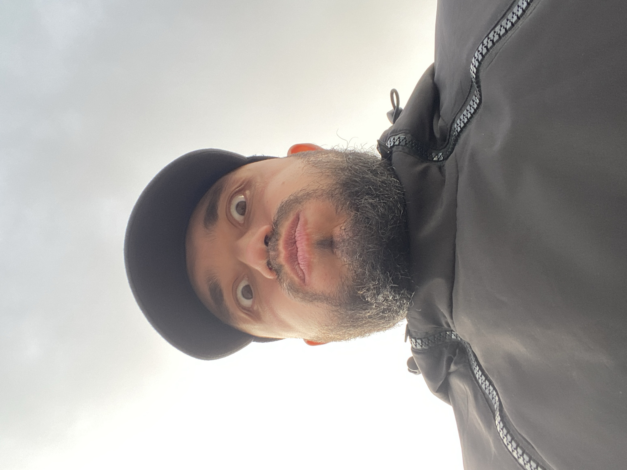 The image displays a close-up, low-angle portrait of a man, rotated 90 degrees clockwise. When viewed upright, the man's face dominates the upper portion of the frame, looking down towards the camera with wide, direct eye contact. He has a dark, full beard and a mustache, and his lips are slightly parted. He is wearing a dark cap, visible on the top left of his head, and a dark jacket or hoodie. The jacket features prominent zig-zag patterned zippers along seams, visible on the right side of the image (which would be his chest/shoulder area when upright). The background is a bright, overcast sky, mostly white with very faint grey cloud variations, indicating an outdoor setting during the daytime. There are no specific landmarks or discernible features in the sky to suggest a precise location, other than the general indication of an outdoor environment. No particular activity is depicted beyond posing for the photograph. The man's expression appears neutral or slightly serious. No visible text is present in the image.
