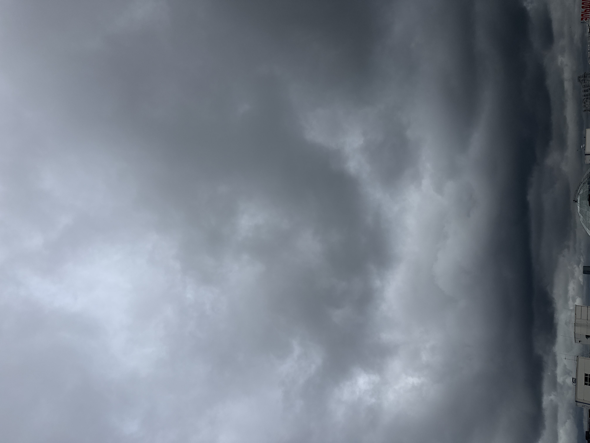 The image shows a dark, overcast sky filled with heavy, grey clouds. In the foreground, the roofs of several buildings are visible, some appearing to be modern office buildings or apartment complexes. The architecture suggests an urban setting. The sky is dominant in the frame, with the clouds creating a dramatic and somber atmosphere. There are no visible people or specific actions taking place. The overall scene is indicative of impending rain or a storm. The image does not contain any explicit clues to its specific location within Sovkhoza imeni Lenina, Russia.