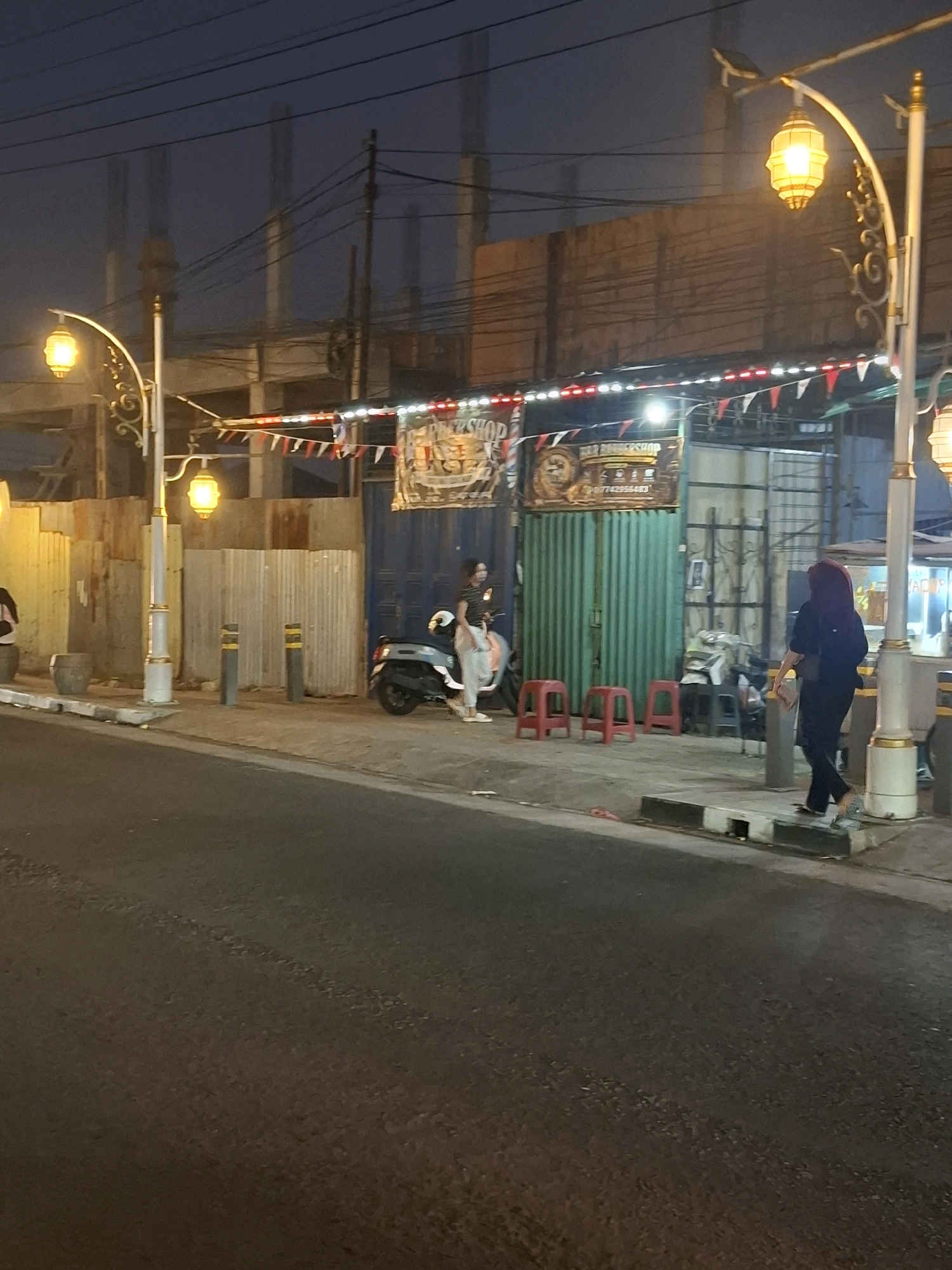 The image captures a street scene at dusk or night in Sayang, Indonesia, likely in a commercial or residential area. Ornate streetlights with glowing yellow lanterns illuminate the pavement and sidewalk. In the background, a building under construction is visible with its skeletal framework and unfinished walls. A corrugated metal fence runs along the side of the street, bordering a row of shops or stalls.
On the sidewalk, several red stools are arranged in front of a green corrugated metal structure, possibly a shop or food stall. A light-colored scooter is parked on the sidewalk. Two women are present. One woman, wearing a white dress and light-colored shoes, is sitting on the scooter. Another woman, wearing a dark headscarf and dark clothing, is walking away from the viewer towards the right of the frame, carrying a light-colored purse. Two dark cylindrical bollards with yellow tops are positioned on the sidewalk. The asphalt road in the foreground shows signs of wear and tear. Decorative bunting with red and white flags is strung above the shops.