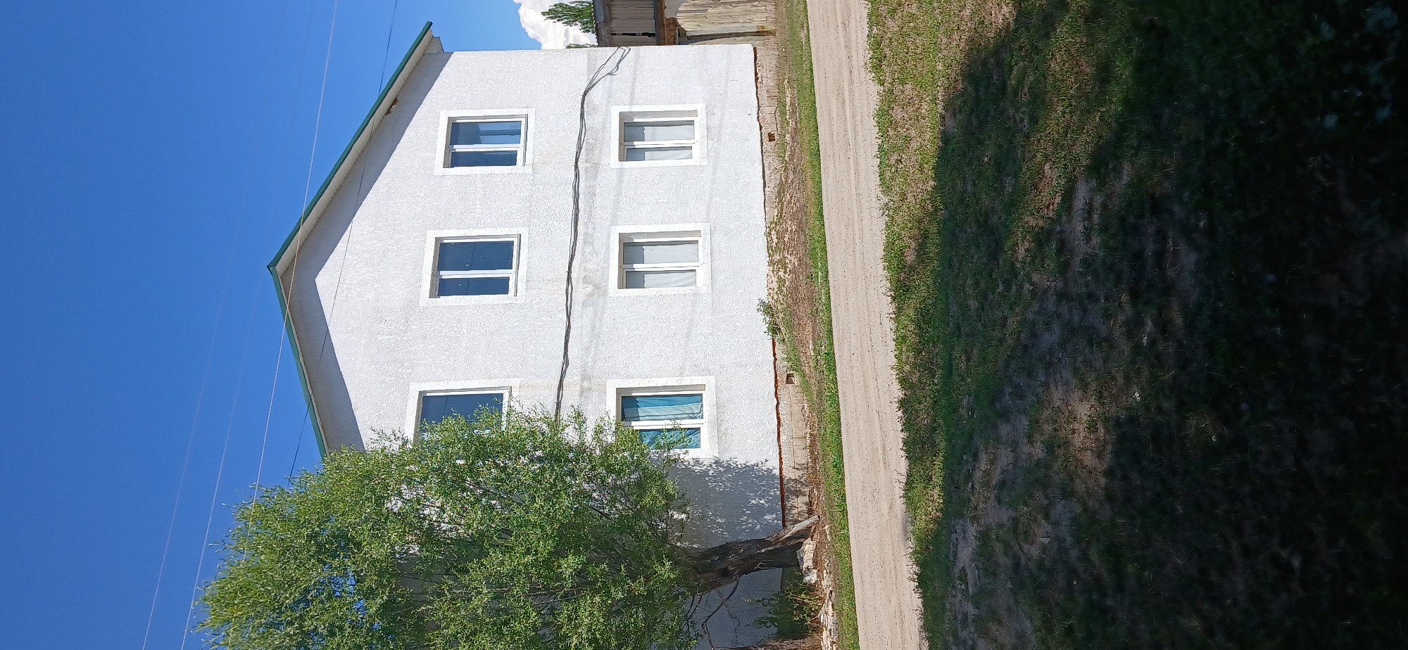 A two-story white stucco house with a green roof is situated in a rural area. The house has six windows on its front facade, three on the upper level and three on the lower level. A large, leafy green tree stands to the left of the house, casting a shadow on the building. A dirt road runs in front of the house, bordered by patchy grass and dirt. In the background, a wooden fence and some distant trees are visible under a clear blue sky. The image was taken in Mihaylovka, Kyrgyzstan.