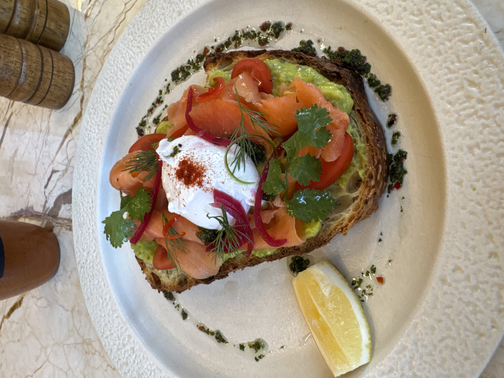 A close-up, overhead shot shows a plate of avocado toast with smoked salmon and a poached egg on a textured white plate. The toast is topped with mashed avocado, thinly sliced smoked salmon, cherry tomato wedges, pickled red onions, and fresh cilantro. A poached egg sits in the center of the toast, sprinkled with paprika. A wedge of lemon is placed to the left of the toast. The plate is drizzled with a green herb sauce. The background consists of a marble tabletop and out-of-focus wooden salt and pepper grinders and a brown mug. The scene appears to be in North Kuta, Indonesia.