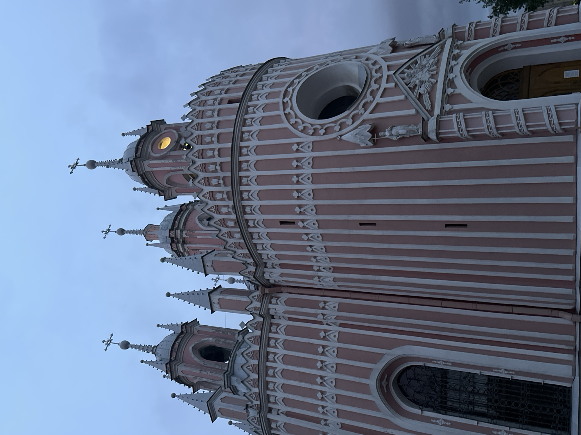 The image shows the Cesme Church in Saint Petersburg, Russia. The church is a religious building with a distinctive pink facade and white architectural details. It features multiple spires topped with crosses, decorative window frames, and a large circular window. The sky is overcast and a dull blue, suggesting it is either early morning or late evening. There are no people or active interactions visible in the scene, only the static architecture of the church and the surrounding overcast environment.