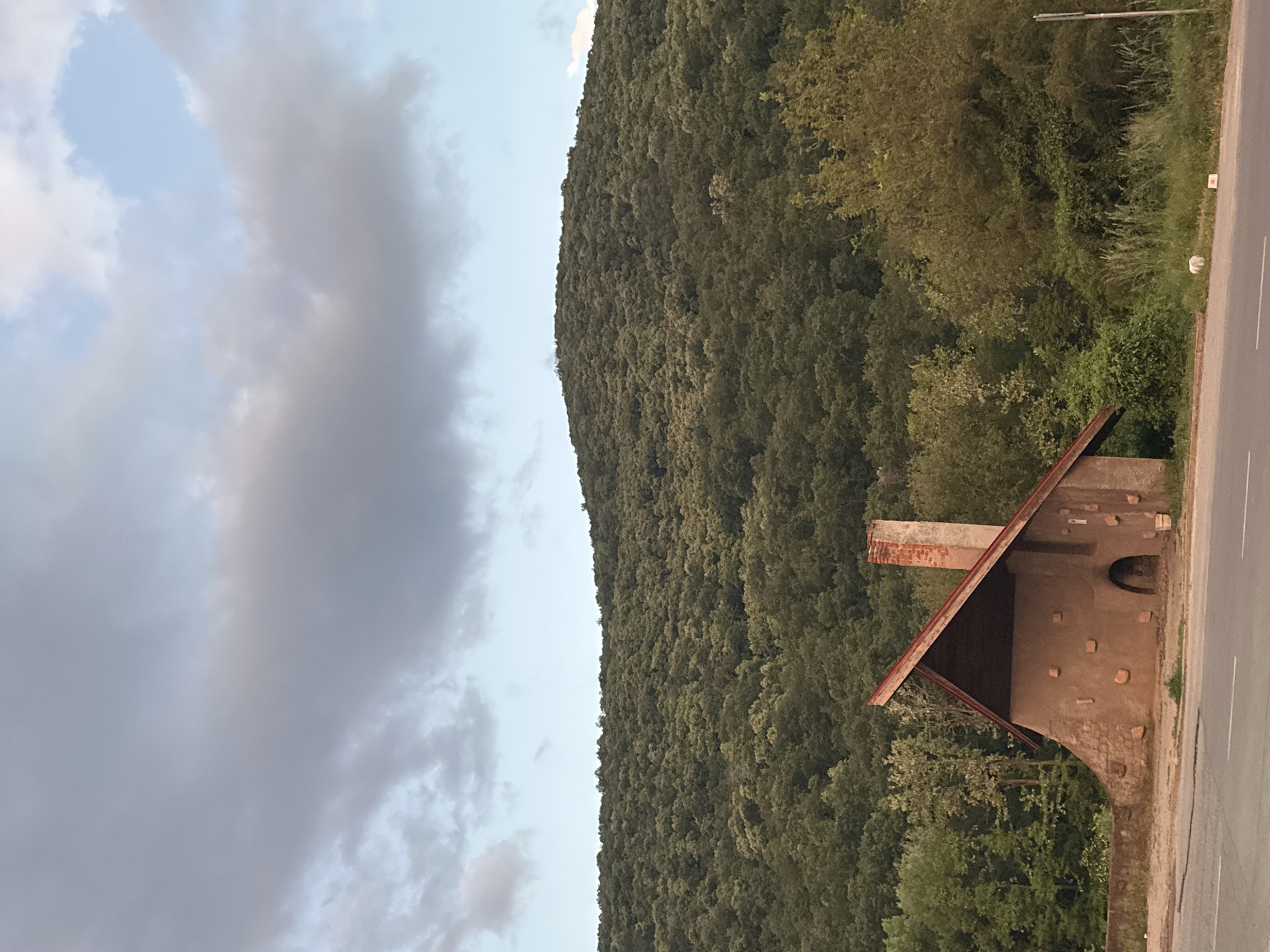 The image shows a roadside building in Pryaporets, Bulgaria, adjacent to a forest-covered hillside. The building is a simple, one-story structure with a slanted roof and a brick chimney. It is constructed from stone or brick and has an arched doorway. A paved road runs alongside the building, and a cloudy sky is visible in the background.