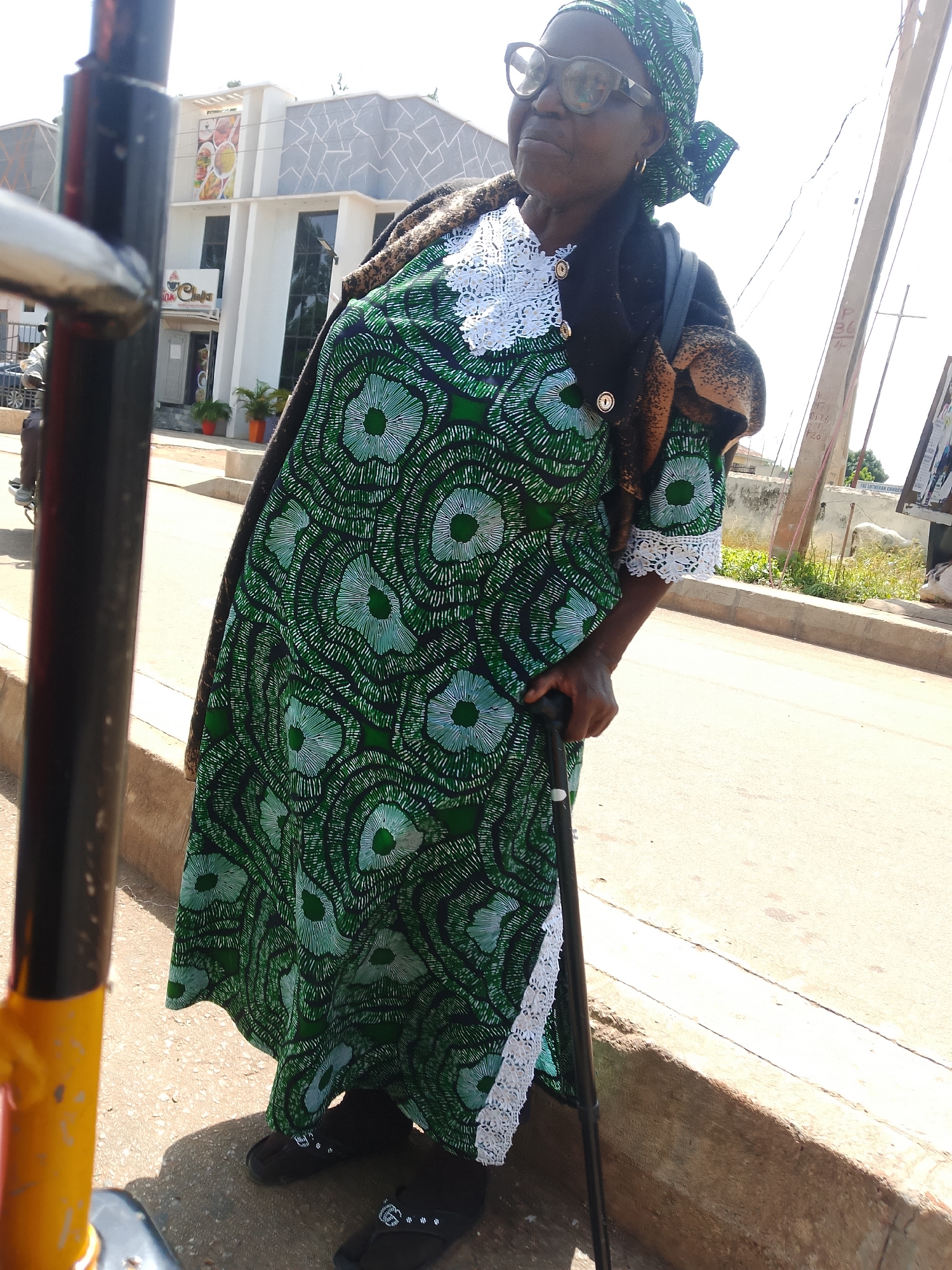 An elderly Black woman wearing a green patterned dress, a headscarf, and glasses stands on a sidewalk holding a cane. She is wearing black flip-flops. In the background, a modern building with a gray patterned facade and tall windows is visible, along with streetlights and a few potted plants. The scene appears to be in Jos, Nigeria, suggested by the architectural style and general ambiance.
