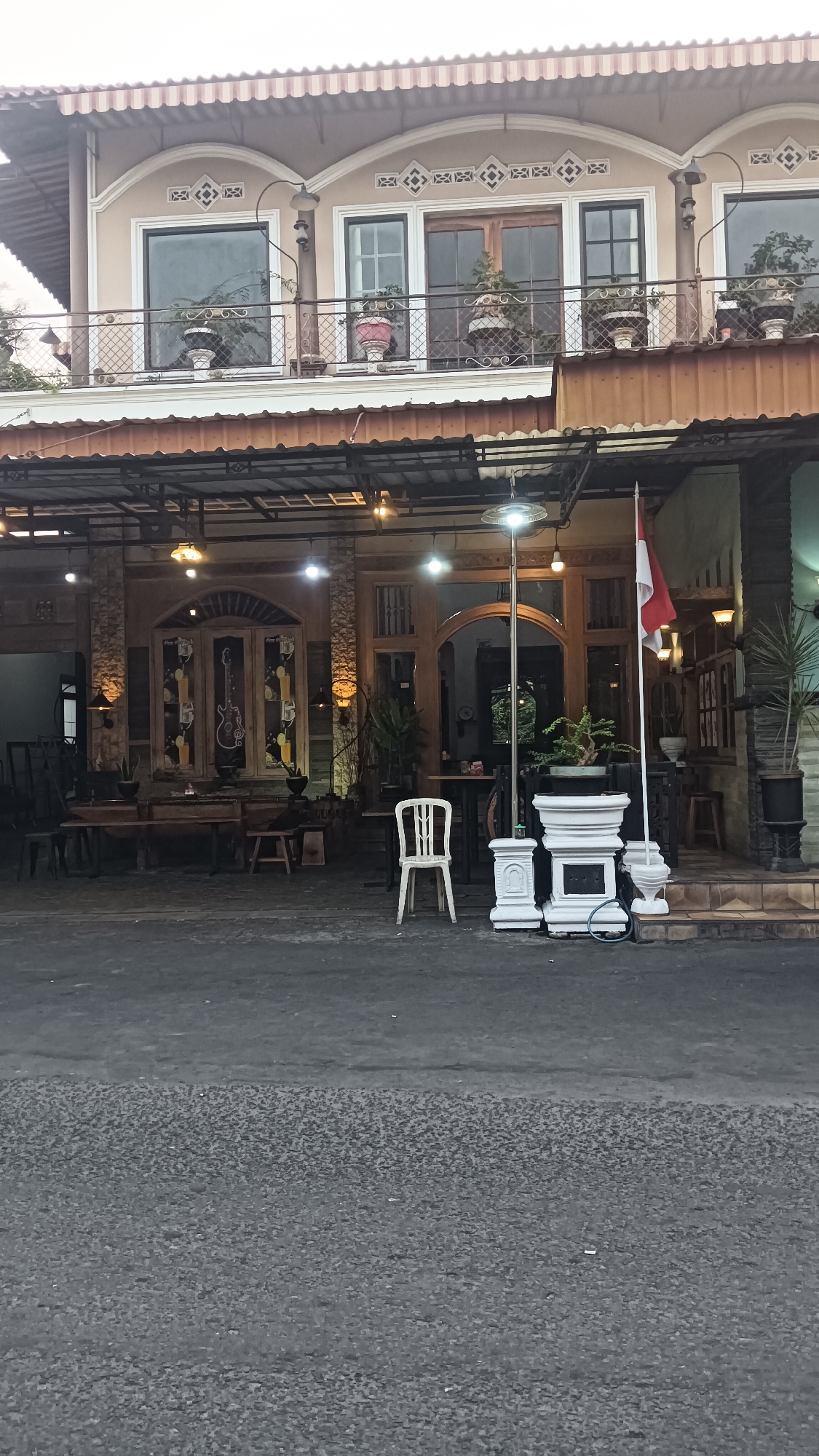 The image shows a storefront in Sragen, Indonesia, likely a cafe or restaurant. The building has two stories with a balcony on the upper level. The storefront itself is made of wood and stone and has an open-air seating area. There are several tables and chairs outside, as well as some decorative items like potted plants and a flag. The street in front of the building is paved with asphalt. The overall impression is one of a casual and inviting establishment.