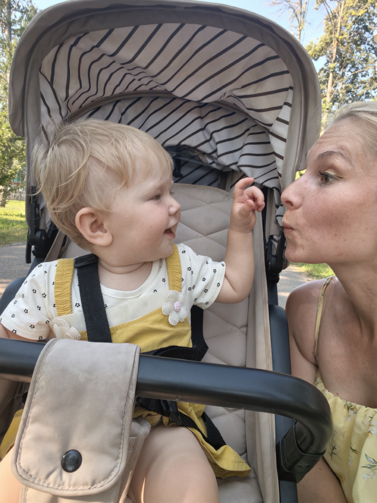 In a medium shot, a woman with blonde hair and yellow eyeshadow is leaning towards a baby in a stroller, puckering her lips as if to kiss the baby. The baby, also with blonde hair, is looking at the woman and reaching out a hand. The baby is wearing a white shirt with small black flowers and a yellow dress. The stroller has a striped interior lining and a tan fabric pouch attached to the handle. The background shows trees and a clear sky, suggesting an outdoor setting, possibly a park or residential area, in Sterlitamak, Russia.