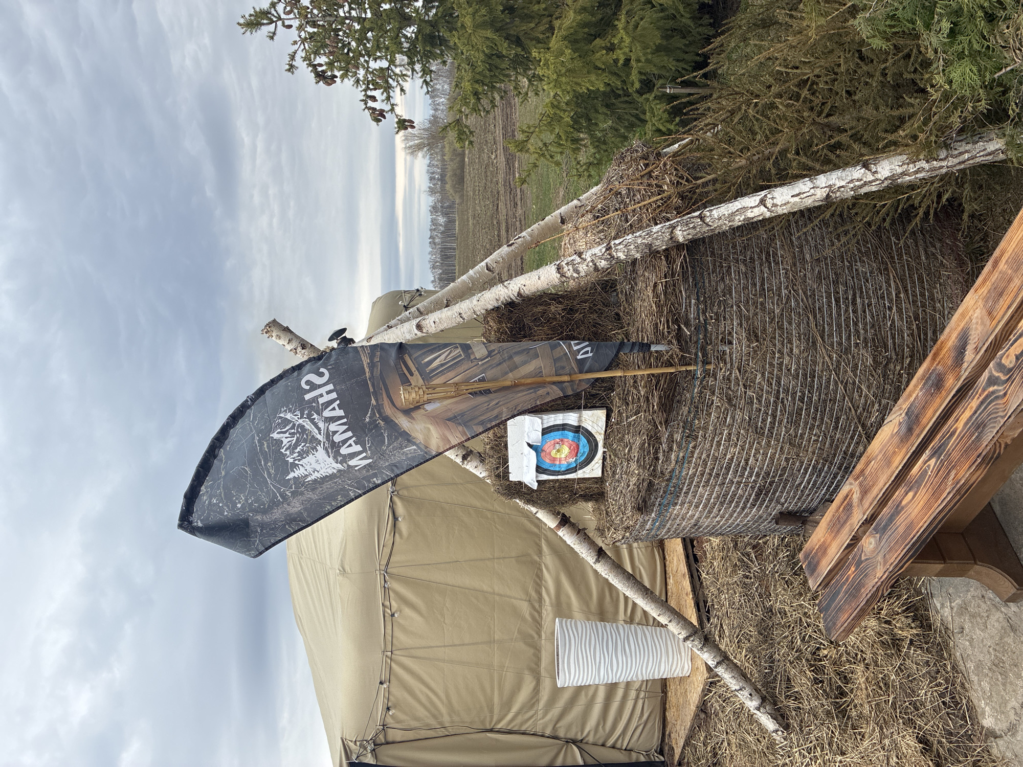 A yurt stands outside, with a bale of hay behind it. The hay bale has a white archery target with red, blue, and yellow rings pinned to it. The target has a torn section. A Yamaha flag is partially visible, and several birch tree branches lean against the yurt and the hay bale. The setting is a field with trees in the background under a cloudy sky. Location: Unknown, Unknown.