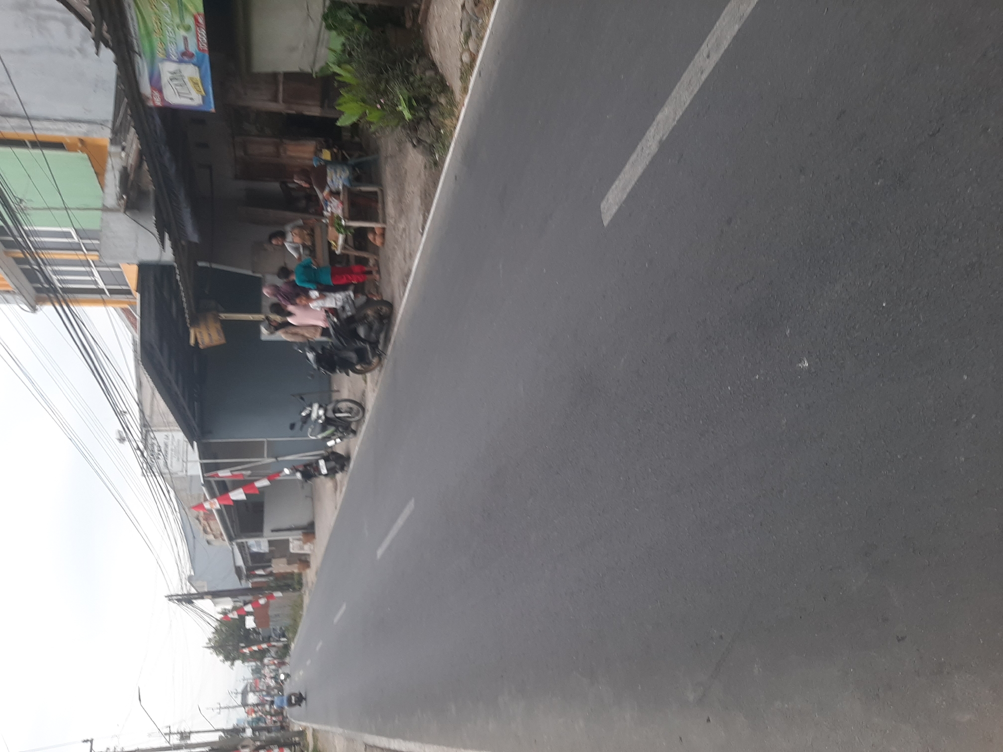 This is a ground-level, medium-angle shot of a paved road in Sumampir, Indonesia, with buildings and people on the right side. The road is a grey asphalt surface with a white line marking the lane. On the right side of the road, there is a row of buildings with corrugated metal roofs and some shops. In front of one shop, a group of people are gathered around a table with food items. Several motorcycles are parked along the side of the road. In the background, there are more buildings and utility poles with numerous wires. The sky is overcast and a few Indonesian flags are visible on poles along the street.