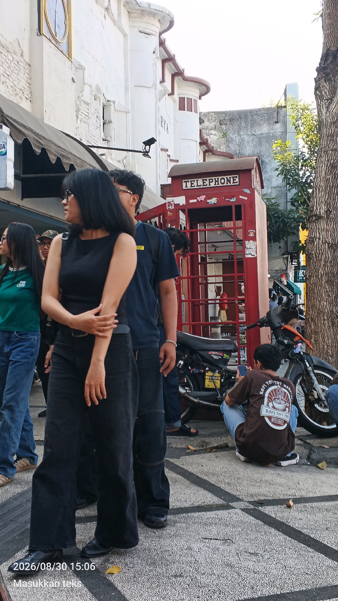 In a bustling urban scene, likely in the city of Malang, Indonesia, a red telephone booth stands prominently as a focal point. A group of individuals are captured in various states of activity. A woman in a black sleeveless top and dark wide-leg pants is in the foreground, her hands crossed over her arm. Behind her, a man in a dark t-shirt walks past. To the left, another woman wearing a green shirt and blue jeans is visible. Further in the background, a young man in a brown t-shirt and light blue jeans is sitting near a motorcycle, engrossed in his phone. The environment suggests a public space, with a tree on the right and buildings in the background, one of which has a clock face. The ground is paved with speckled concrete, marked with dark lines. The overall atmosphere appears to be a casual outdoor gathering or a busy street scene.