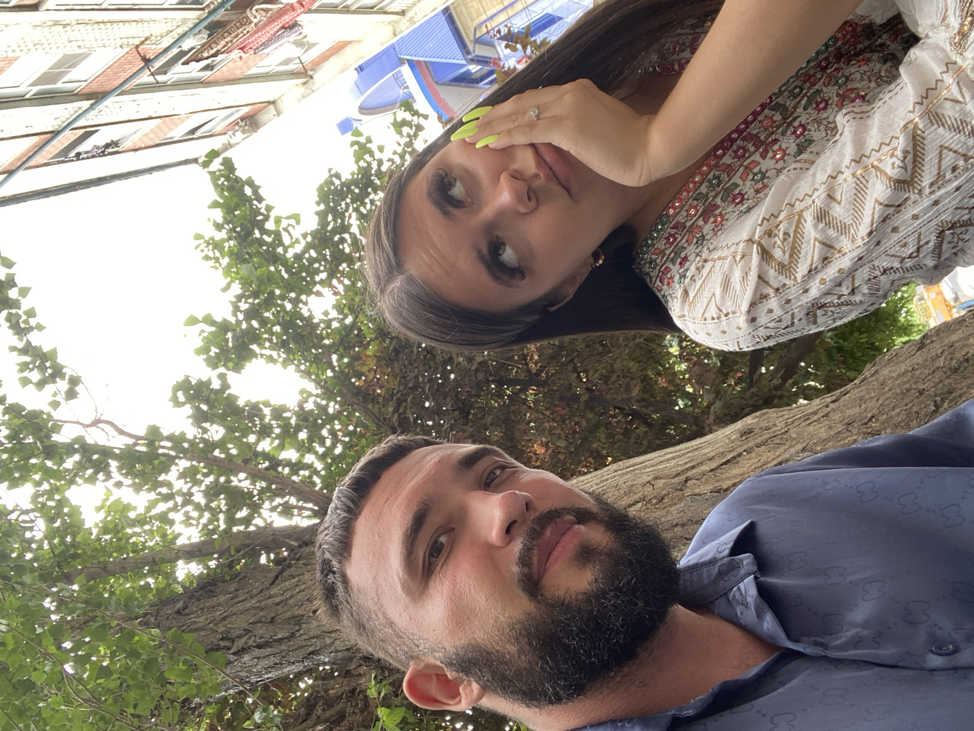 The media file shows a selfie taken outdoors, with a man and a woman as the subjects. The woman is in the upper portion of the image, leaning her head on her hand and looking at the camera. She has long dark hair, bright neon yellow nail polish, and is wearing a white garment with colorful embroidered patterns. A ring is visible on her hand. The man is in the lower portion of the image, also looking at the camera. He has a beard and is wearing a dark blue shirt with a subtle repeating pattern.
They appear to be near a large tree, with its textured bark and green leaves visible in the background. Above the tree, there are hints of buildings and a blue structure, suggesting an urban or semi-urban setting. The lighting suggests it is daytime. The overall mood appears casual and perhaps playful, given the selfie format and the woman's expression. No visible text is present in the image. The location context provided is Derbent, Russia.