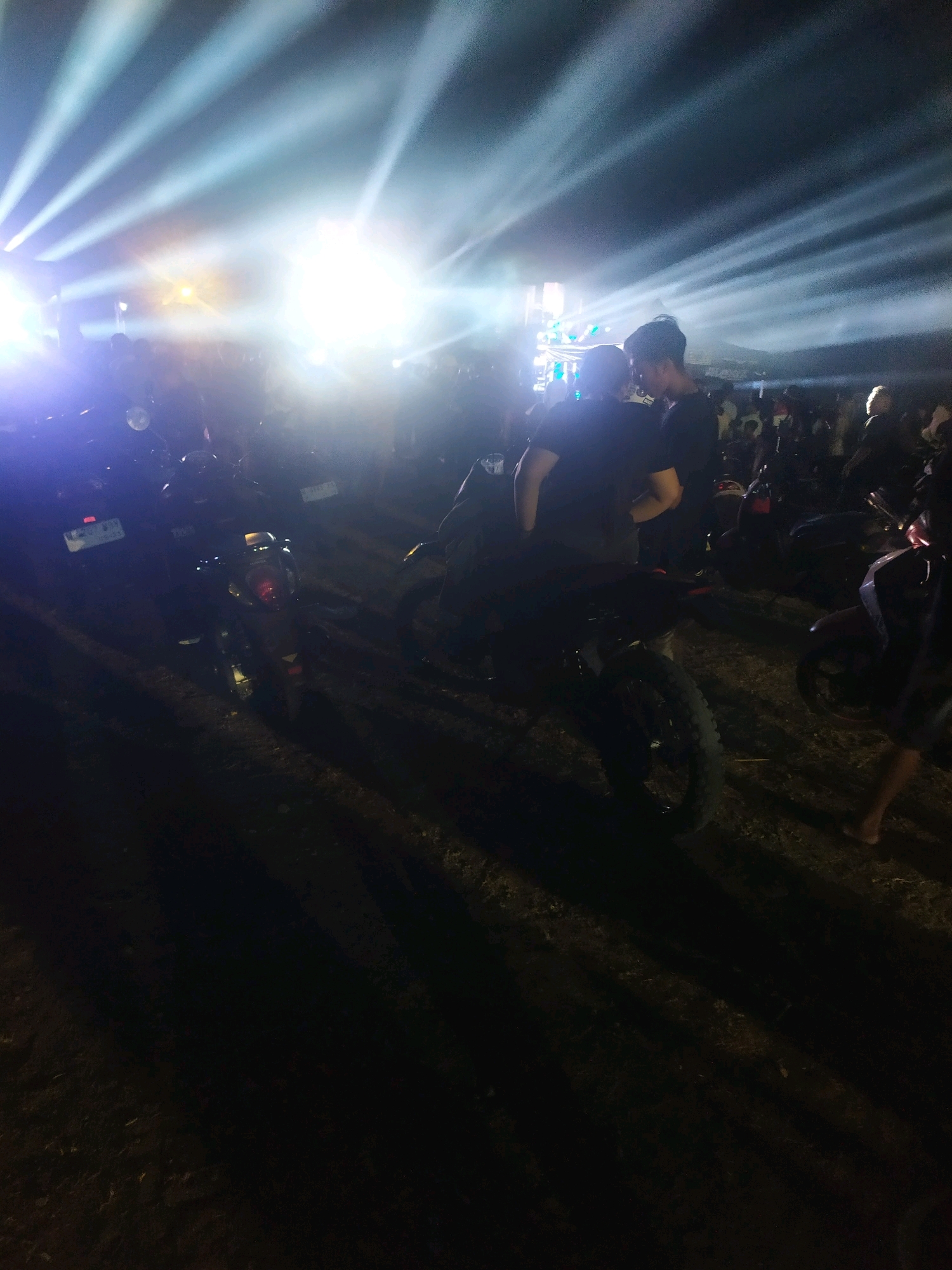 This image depicts an outdoor night scene, likely at an event in Gempol, Indonesia, illuminated by bright stage lights that create dramatic beams across the dark sky. In the foreground, several motorcycles are parked on a dirt or gravel surface. Two individuals, appearing to be young men, are in the center of the frame, facing each other. One is wearing a black t-shirt and is partially obscured by the other. The second man is also in a black t-shirt, facing left, with his head bowed as if in conversation or a tender moment. They are standing close together amidst the parked motorcycles. Behind them, a crowd of people is visible, also silhouetted against the bright lights. The overall atmosphere suggests a lively gathering or concert. The lighting creates a stark contrast between the brightly lit background and the darker foreground, with shadows cast by the motorcycles and people.