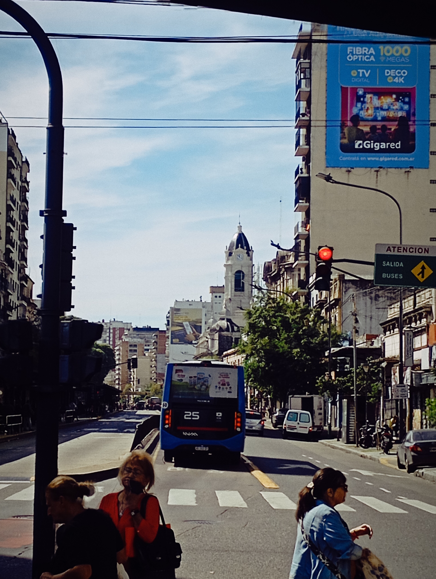 The image captures a street scene in Buenos Aires, Argentina. The viewer looks from a street corner towards a line of buildings with diverse architecture; in the middle is a building with a clock tower. In the immediate foreground, three women are on the corner waiting to cross the street; two are on the left. A red traffic light is lit, stopping traffic, including a blue bus with the number 25. A large advertising billboard is on one of the buildings, promoting fiber optic internet services. The sky is partly cloudy.