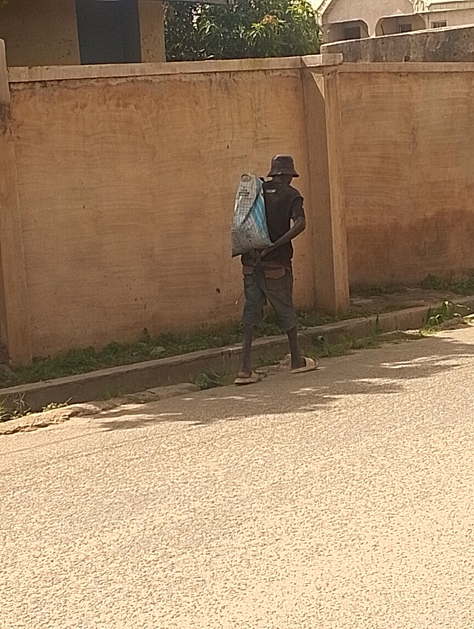 A person wearing a dark bucket hat and a dark t-shirt is walking on a paved road. They are carrying a large, light-colored bag slung over their left shoulder. The bag appears to be made of plastic and has blue and white stripes. The person is wearing dark shorts and light-colored sandals. The scene appears to be outdoors, with a textured brown wall to the left and some greenery and a concrete drainage ditch in the foreground. The sun is casting shadows on the road, suggesting it is daytime. The overall setting is a street in what appears to be a residential or commercial area, possibly in Jos, Nigeria, given the context. The person's posture and the way they are carrying the bag suggest they are transporting goods. There is no visible text in the image.
