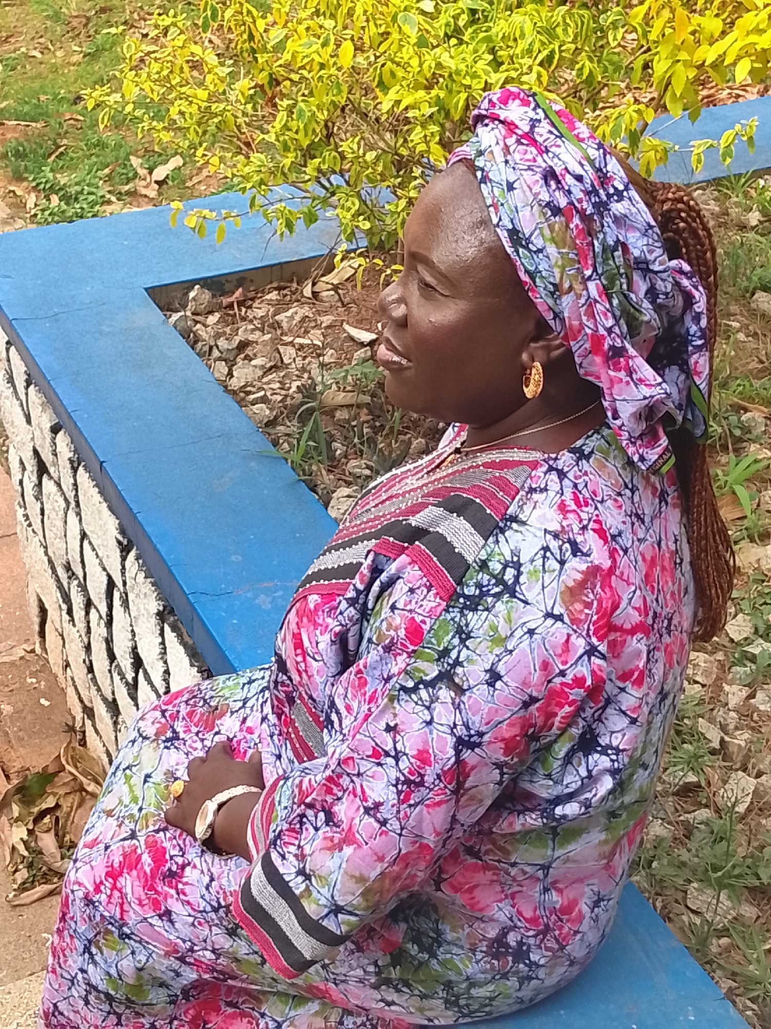 The image features a woman seated outdoors in a bright, natural setting, likely in Zawan, Nigeria. The woman, with dark skin, is shown in profile, looking towards the left side of the frame with a gentle smile, conveying a sense of calmness and contentment. Her hair is styled in braids, one of which is visible over her right shoulder.
She is dressed in vibrant, traditional attire, consisting of a colorful headscarf and a matching long-sleeved dress. The fabric of both garments features a rich, multi-toned pattern with shades of pink, purple, white, and green, characterized by an abstract or floral design. The dress has an elaborate neckline and shoulder area, detailed with black, white, and pink stripes, possibly embroidered or beaded. She accessorizes with golden hoop earrings and a light-colored bracelet or watch on her left wrist.
The woman is seated on a low, blue-painted ledge or wall. Behind her, another section of this blue structure forms a corner. Beneath the blue surface, there is a stone wall with rough-hewn, white-painted stones. The background includes earthy ground with scattered small rocks and sparse green vegetation. Further back, a bush with bright yellow and green variegated foliage suggests a sunny, outdoor environment. The lighting indicates it is daytime. There is no discernible text visible in the image.