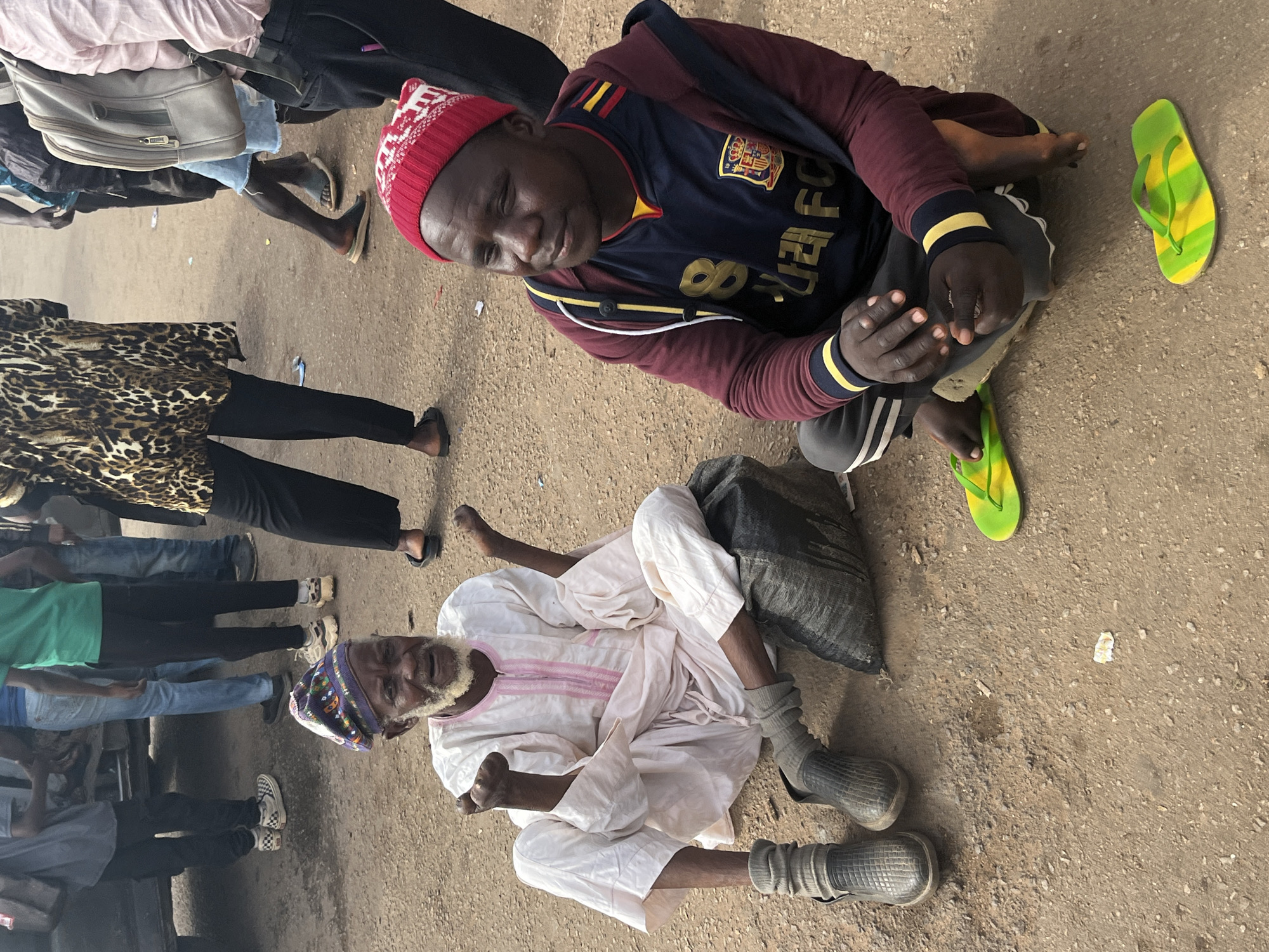 In Gura Top, Nigeria, two men are seated on a dirt ground. The man on the left, an elderly individual with a white beard and a patterned beanie, wears a white robe and gray socks with black slip-on shoes. He sits on a black plastic bag, with his hands raised and palms open. The man on the right, younger, wears a red beanie, a dark blue and yellow jersey, and a maroon hoodie with white stripes on the sleeves. He sits with his legs crossed, and a pair of yellow and green flip-flops are visible next to him. In the background, several people are standing, with a vehicle partially visible. The environment appears to be an outdoor, possibly dusty, urban setting.