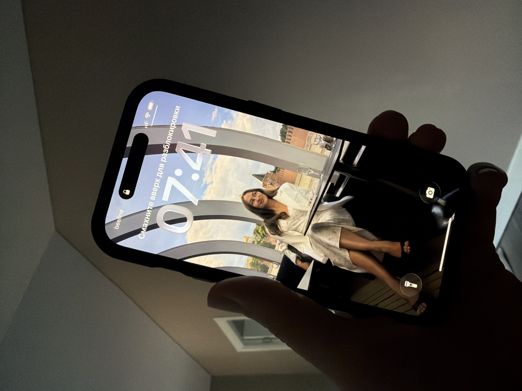 A person's hand is holding a smartphone. The smartphone's screen displays a lock screen with the time 07:41. The background wallpaper on the phone is a photo of a woman sitting at a table, with a city skyline visible through a large window. The city skyline appears to be that of Malinovka, Russia, featuring historic architecture. The phone has icons for a flashlight and a camera at the bottom of the screen.