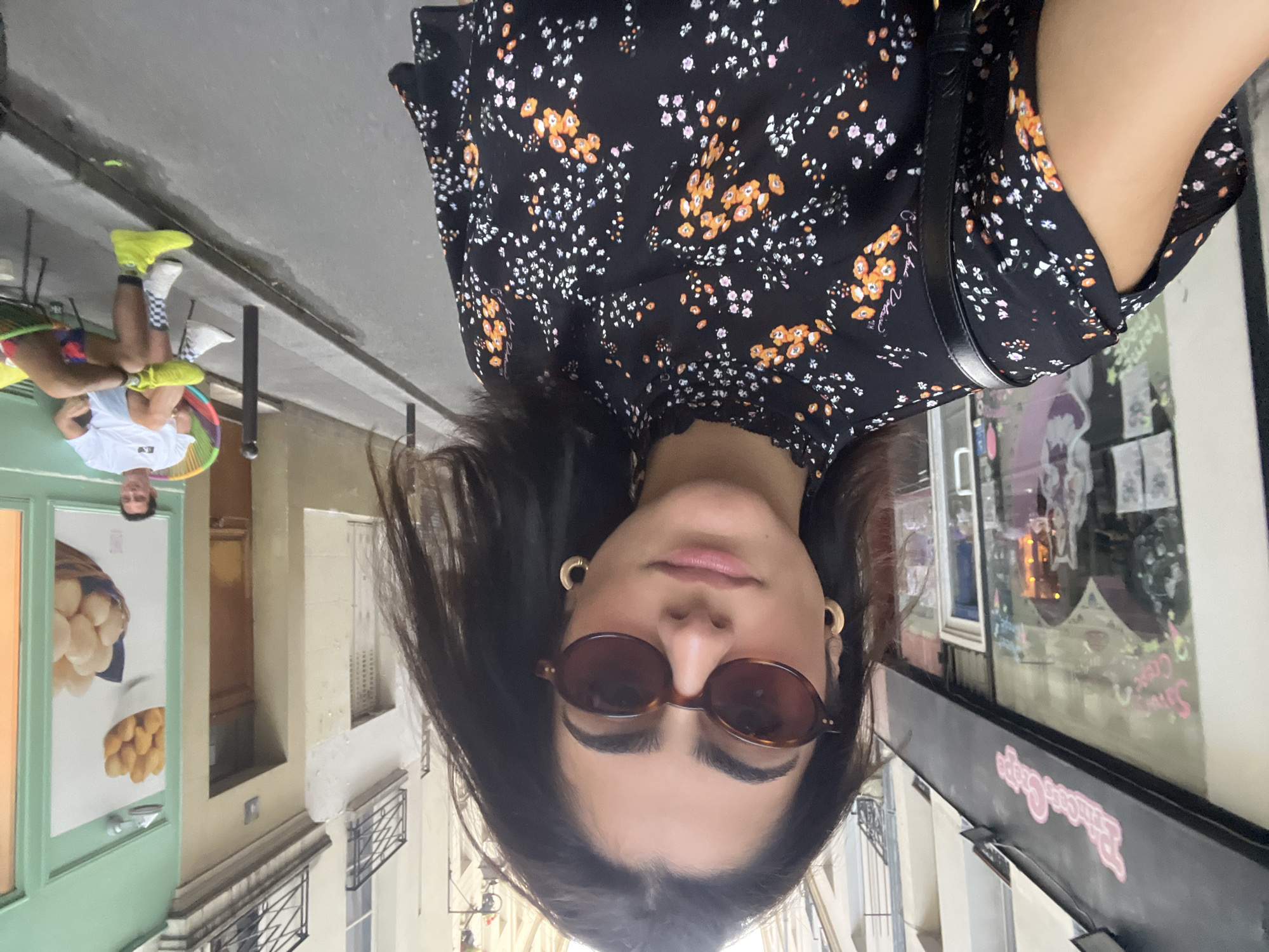 In Paris, France, a woman with dark hair and sunglasses takes a selfie on a narrow street. She is wearing a black dress with a floral print and gold hoop earrings. To her left, a shop called "Princess Crepe" is visible with colorful decorations in the window. Further down the street, a man with short brown hair sits in a chair outside a building with a mint green facade. He is wearing a white t-shirt, light blue shorts, and bright yellow sneakers. He is looking towards the camera and smiling. The street is paved with cobblestones, and buildings with balconies line both sides of the street. The overall scene suggests a casual, urban setting in Paris.