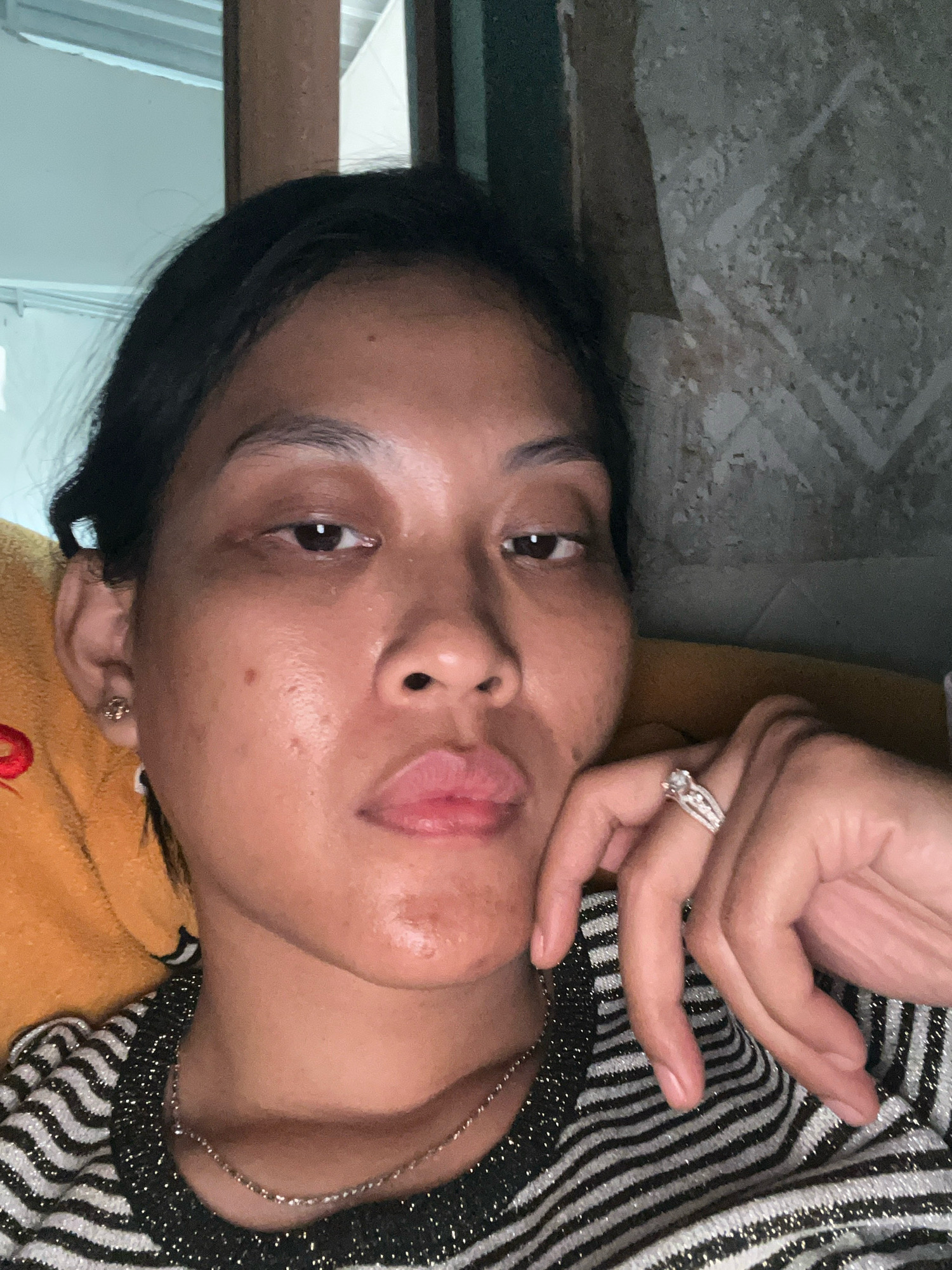 This is a close-up, selfie-style portrait of a young woman in what appears to be an indoor setting. She has dark hair pulled back, exposing her face and ears. She is wearing a striped shirt with a sparkly trim and a delicate necklace. Her right hand is resting on her cheek, and she is wearing a ring with multiple stones on her ring finger. The background suggests a rustic or unfinished room, with textured walls and what looks like wooden beams. The lighting is soft, creating a slightly moody atmosphere. The location of Kecamatan Pati, Indonesia, is not visually discernible from the provided image.