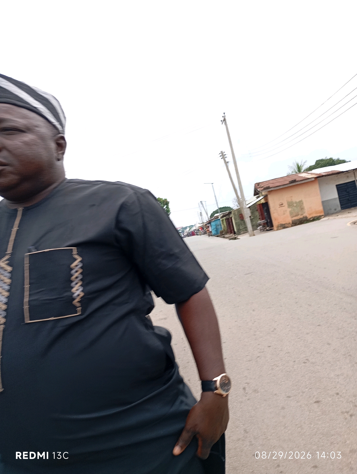 A medium-shot, eye-level, outdoor photograph taken in Shendam, Nigeria, shows a man walking on a paved street. The man, from the waist up and facing left, wears a black traditional outfit with intricate gold and brown embroidery on the left chest pocket and down the front opening. He also wears a black and white striped cap and a gold-faced wristwatch on his left wrist. The street is lined with buildings on the right, featuring corrugated metal roofs and various colored walls, some appearing worn. Electrical poles and wires are visible in the background, and the sky is overcast. The bottom left corner of the image displays "REDMI 13C," and the bottom right corner shows the date and time "08/29/2026 14:03."