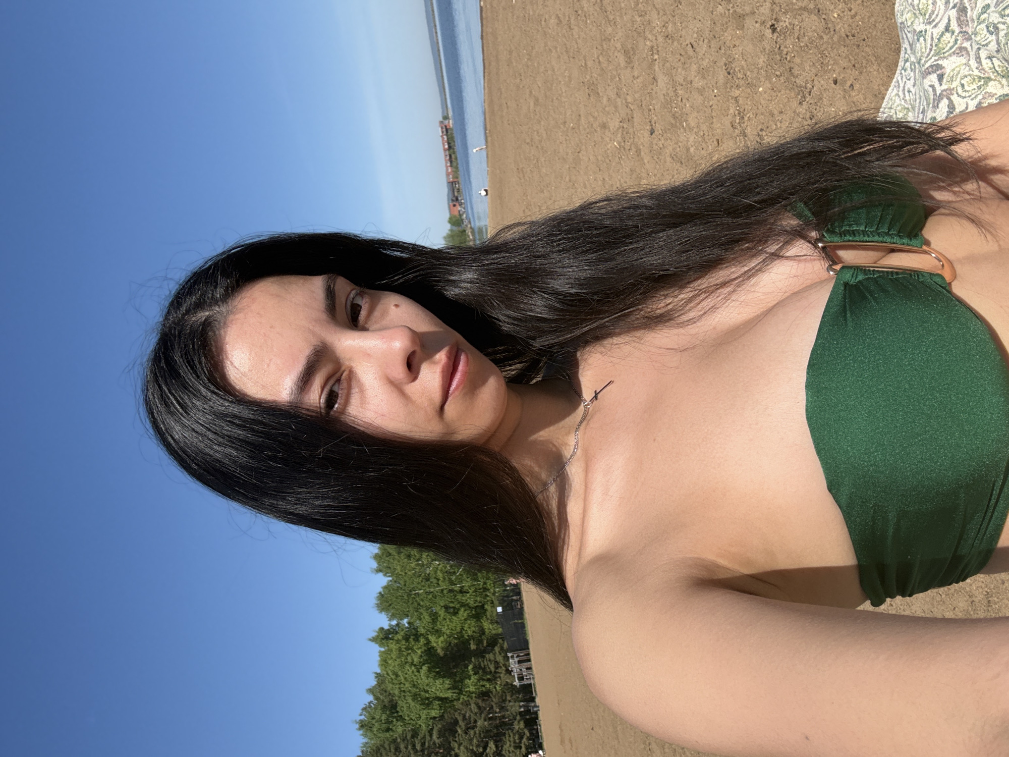 A woman lies on a beach in Chaikovsky, Russia. She has long, dark hair, light skin, and is wearing a green bikini. The beach is sandy, and there is water in the background. Trees can be seen in the background. The sky is blue and sunny. A silver cross necklace can be seen on her neck. A blanket is seen to her right.
