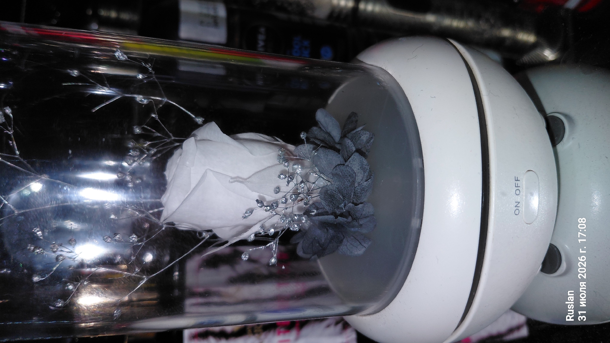 The image shows a preserved white rose inside a glass dome, which sits on a white base with an "ON/OFF" button. The base is part of a device, possibly a humidifier or diffuser, which is reflecting light. In the background, blurred bottles and other objects are visible, suggesting an indoor setting. Text at the bottom of the image indicates the date "31 июля 2026 г. 17:08" and the name "Ruslan". The city of Oleksandriia, Ukraine, is mentioned, but its presence is not visually confirmed within the image itself. The scene appears to be a still life arrangement in a domestic or retail environment.