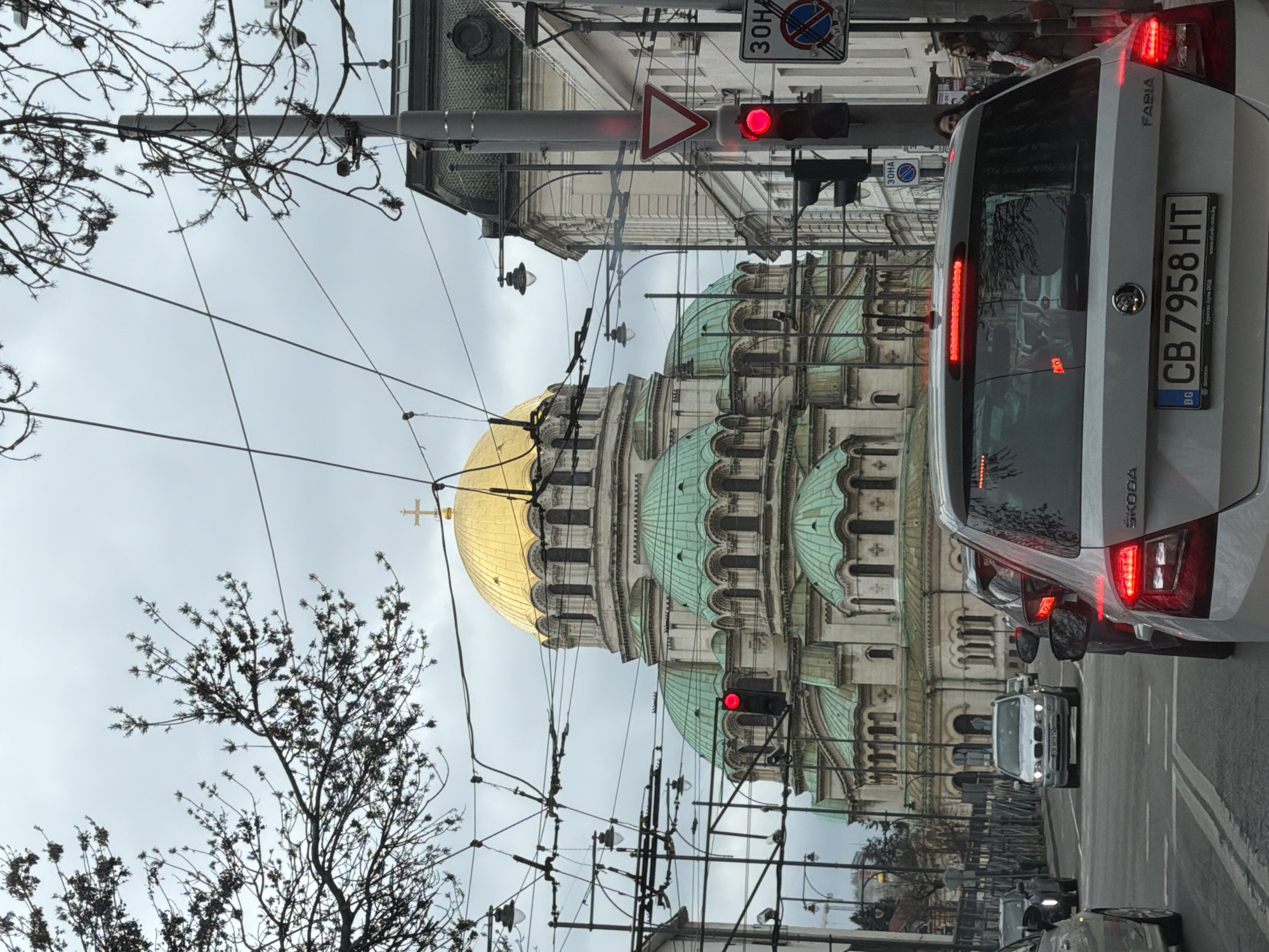 The image depicts the Alexander Nevsky Cathedral in Sofia, Bulgaria, taken from a street level. A silver Skoda Fabia with Bulgarian license plate "CB 7958 HT" is prominently featured in the foreground, its brake lights illuminated. A traffic light above displays a red signal. A road sign with a "30HA" restriction is also visible. Bare trees and power lines are present on the left. Several other cars are on the street. The sky is overcast.