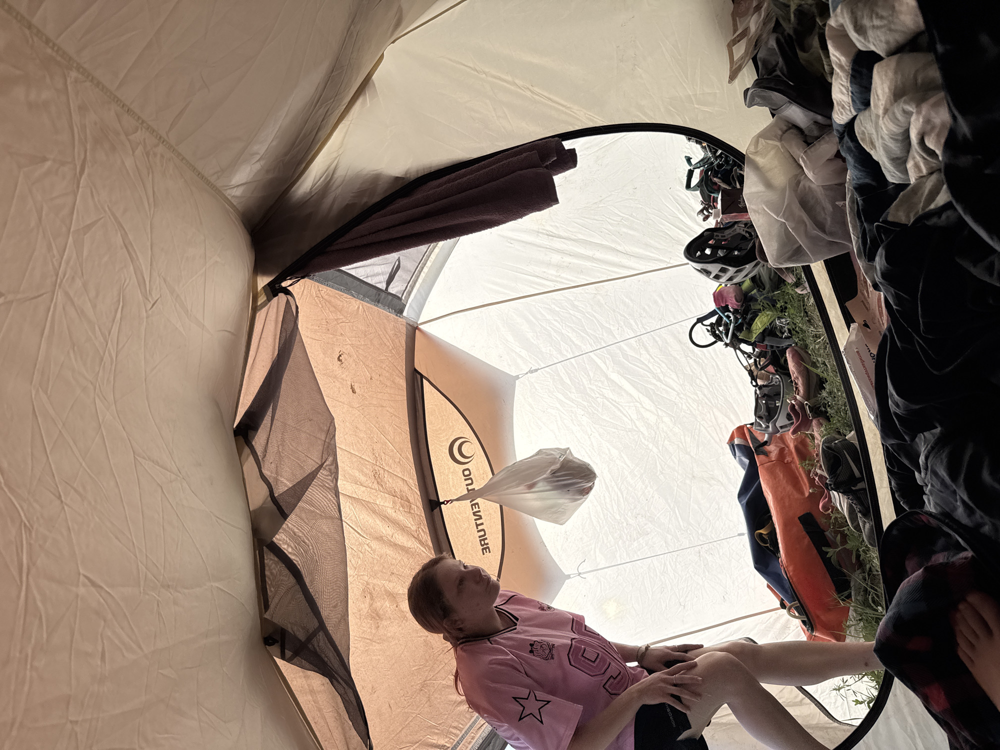 A low-angle, distorted wide shot shows the interior of a tent. A woman sits within, wearing a pink jersey with black shorts. Her legs extend toward the tent opening. A plastic bag hangs from the tent's interior, attached to the seam. Outside the tent opening is a pile of gear including clothing, shoes, and a helmet. The tent is situated on a patch of grass. The location and city of Khvalynsk, Russia cannot be determined from the image.