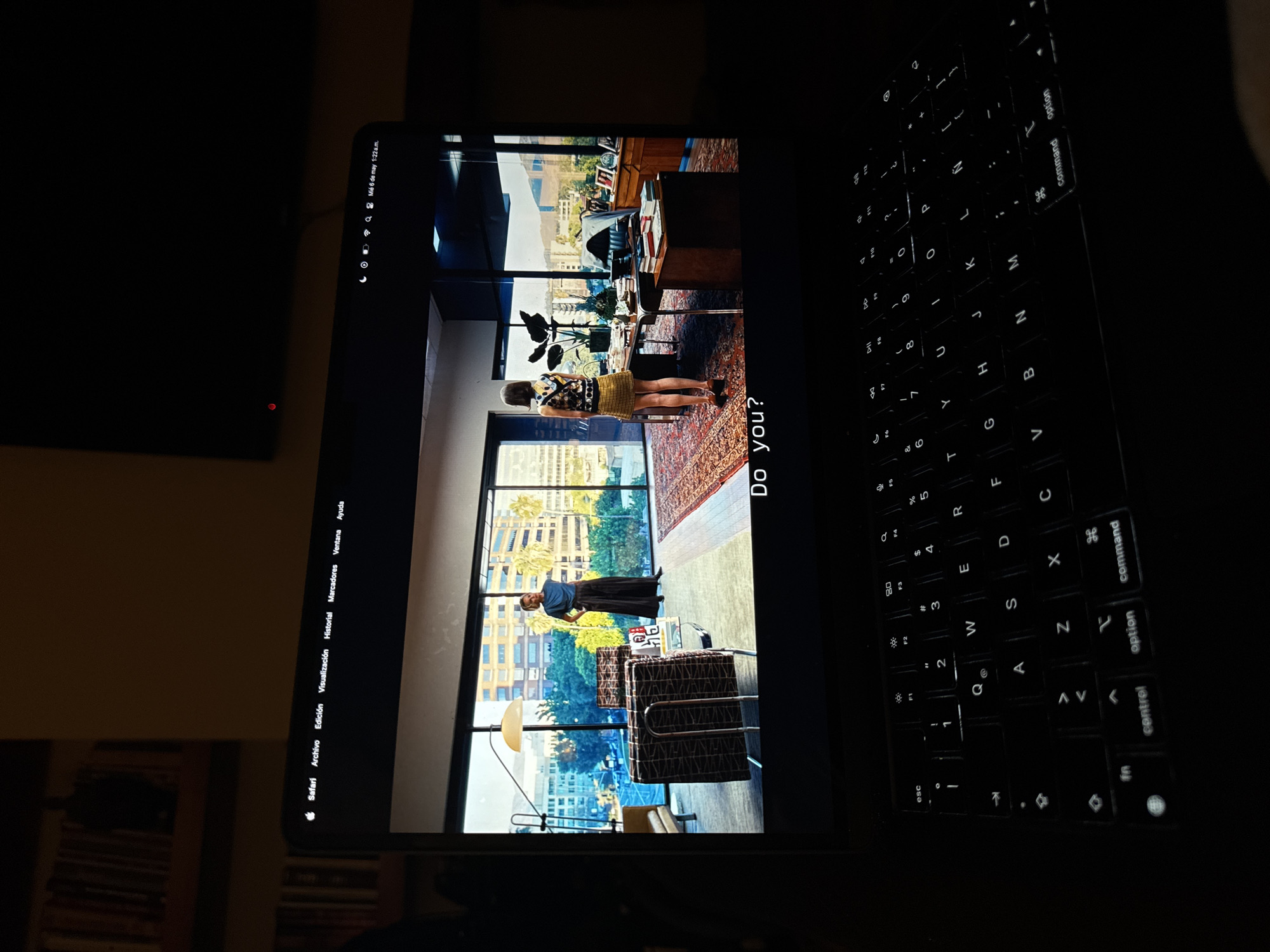 The image shows a MacBook screen displaying a photograph. The photo is an interior shot of an office space featuring two individuals. One person stands near a window with a view of Mexico City's skyline. The other person is in the back, facing toward the window with a yellow skirt and a black vest. The office is decorated with a red oriental rug and some dark furniture. The MacBook interface includes the Safari browser and macOS menu bar. The photograph has text superimposed that reads "Do you?". The keyboard of the MacBook is visible in the right side of the image.