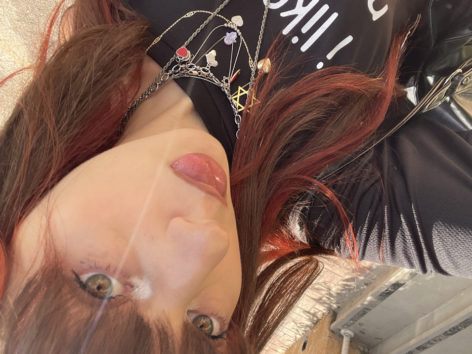 A close-up, angled portrait of an individual, likely female, captured outdoors in Raanana, Israel. The subject has dark brown hair with red highlights and bangs, green/hazel eyes with visible makeup, and glossy reddish-pink lips. A diagonal lens flare crosses the upper left portion of the face. She is wearing a black textured shirt with partially visible white text reading "i like" on the right side of the chest. Multiple layered necklaces adorn her neck, including a prominent dark-toned chain with charms such as a gold Star of David, a red heart, a white cloud, and purple shapes. A thinner gold-colored chain is also present. A dark strap, possibly from a bag, is visible on the lower right. The background is blurred, showing hints of light-colored structures on the left and a bright, textured surface on the upper right, indicative of strong sunlight.