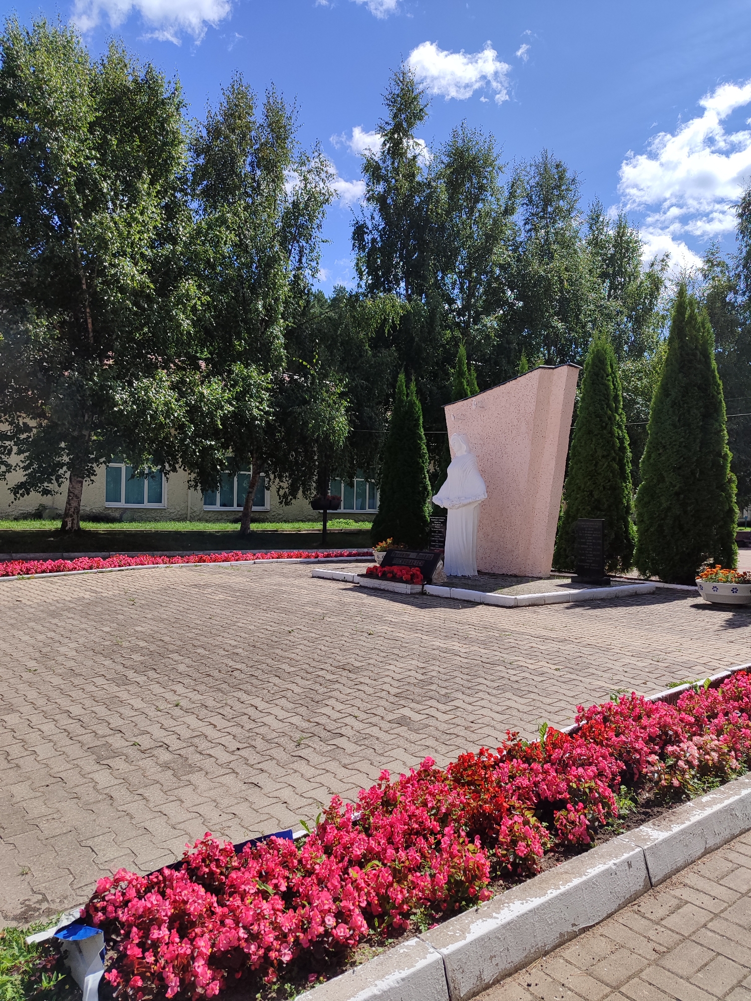 This is a full outdoor shot of a public memorial in Unknown, Unknown, on a bright, sunny day with a blue sky and scattered white clouds. In the foreground, a paved area of interlocking beige bricks is bordered by a curb and a vibrant flower bed of pink and red begonias. To the left and right of the paved area, more flower beds are visible. In the background, a light-colored building with multiple windows is partially obscured by large, leafy green trees, possibly birch trees due to their light bark. The central feature of the scene is a memorial monument composed of a large, angled pink slab behind a white statue of a robed figure. Several tall, conical evergreen trees stand behind and to the sides of the monument. Two black plaques are affixed to the base of the memorial. A trash receptacle is visible to the right of the monument. The overall scene depicts a well-maintained outdoor space.