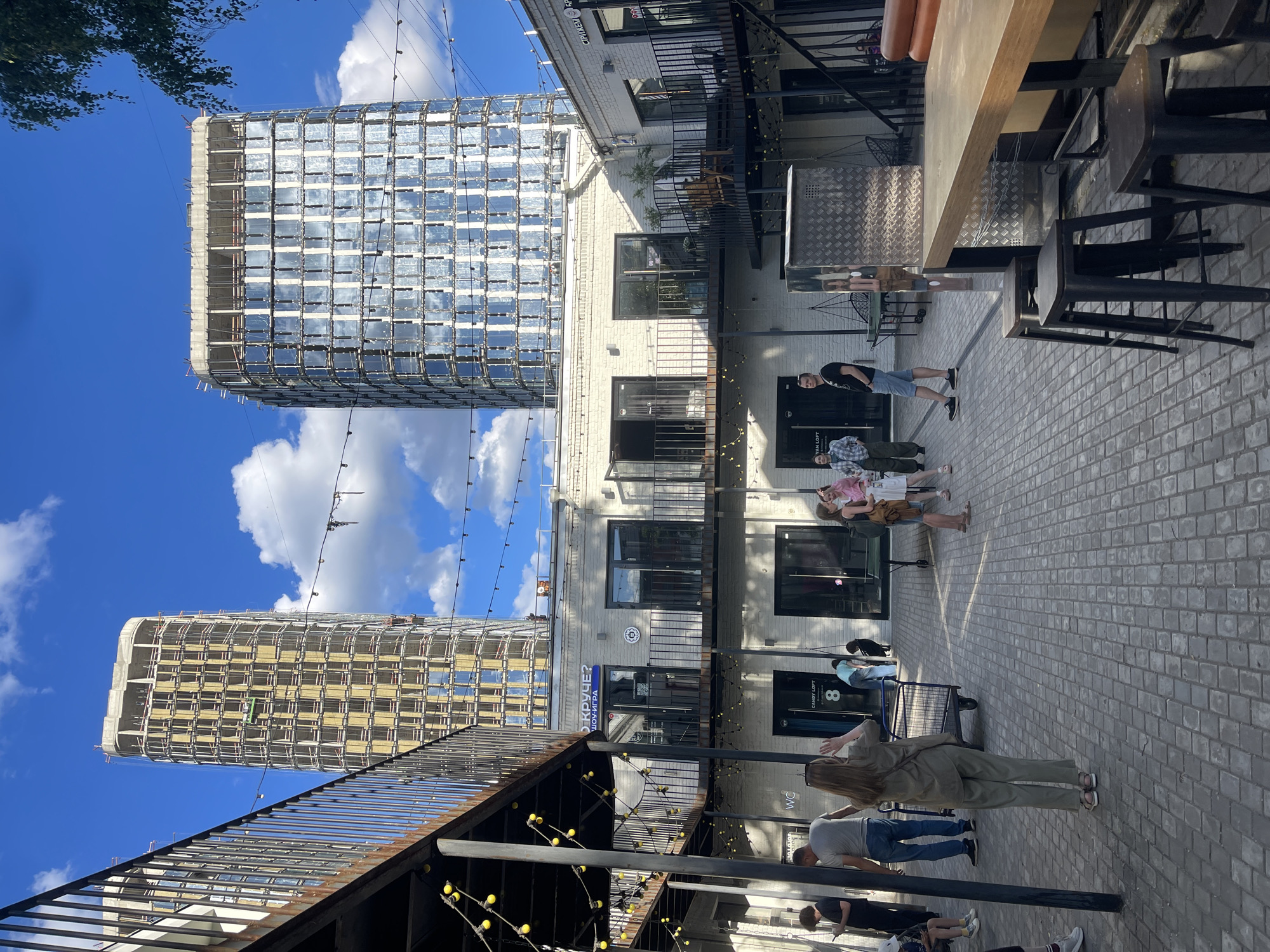 The image shows an outdoor scene in Moscow, Russia, on a sunny day with a blue sky and scattered clouds. The perspective is from a lower level looking upwards and across a courtyard area. In the background, two modern, tall buildings are under construction. One building is mostly covered in glass, reflecting the sky, while the other reveals its concrete structure with wooden scaffolding.
In the foreground and midground, a paved courtyard area is lined with low-rise, light-colored brick buildings. These buildings appear to house shops or businesses, with signage visible, including "CANDY LOFT" and "WC". Several people are scattered throughout the courtyard, some walking, some standing in small groups, suggesting a casual daytime activity. A man in a black t-shirt and blue shorts is standing near a table and chairs, looking towards the camera. A group of three women are standing together, and another person is walking a dog. In the lower left, a metal staircase with decorative string lights is visible, leading to an upper level that is not fully shown. The overall atmosphere seems relaxed and urban, with a mix of ongoing construction and established commercial spaces.