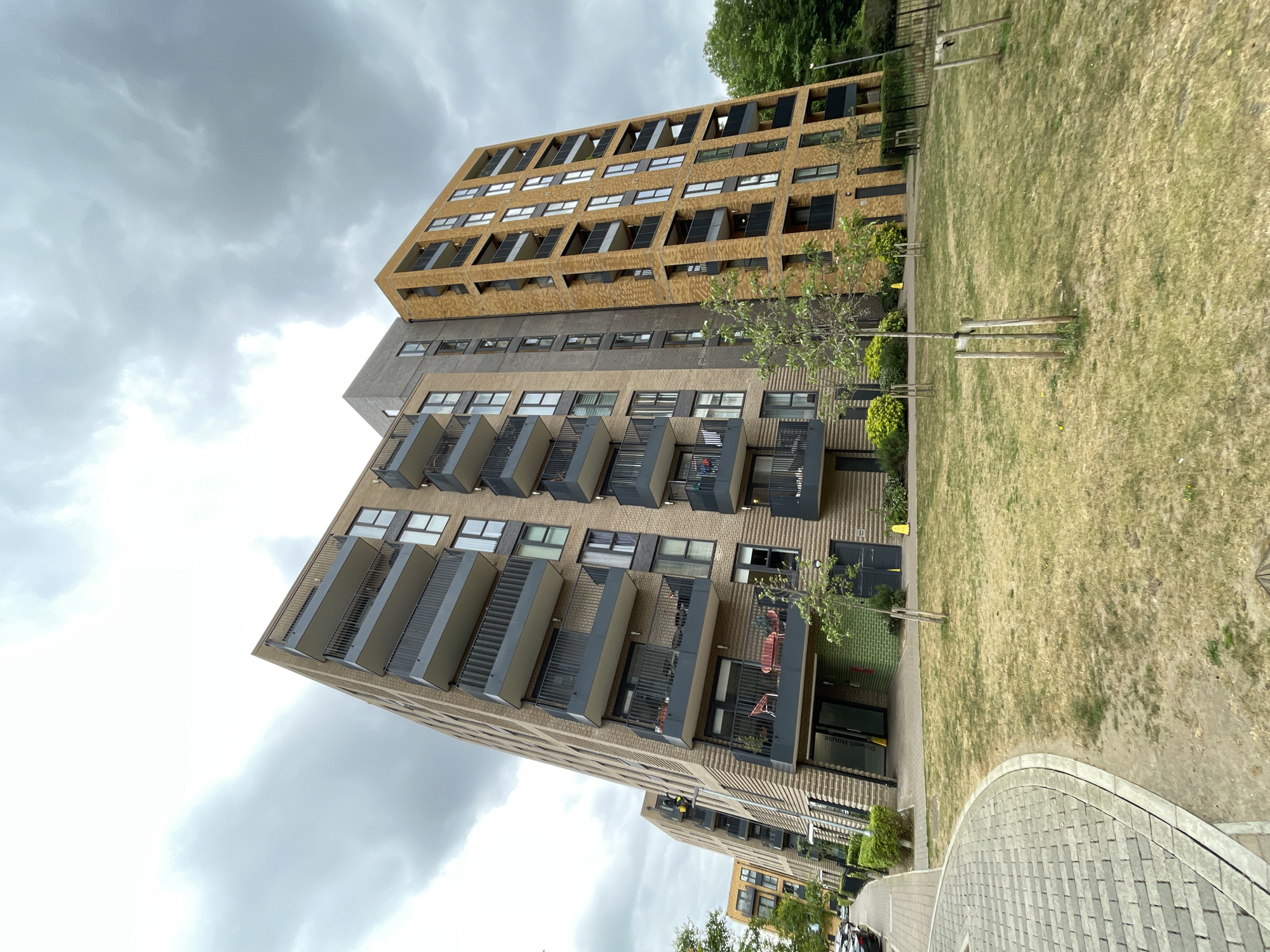 The image shows a modern residential building in London, United Kingdom. The building has a combination of brick and concrete facades, with multiple floors and balconies. There is a grassy area in front of the building, with young trees planted along the walkway. The sky is cloudy and overcast. A paved walkway is visible in the foreground. The architecture is contemporary, and the scene appears to be a residential area within the city.