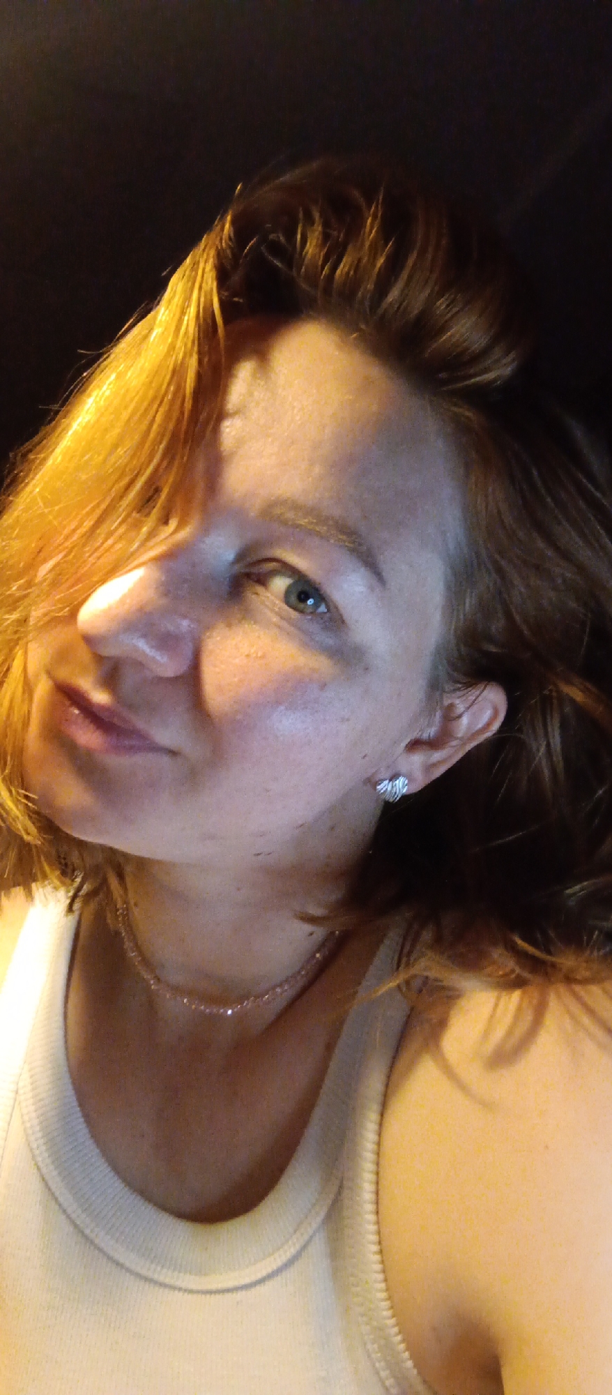A medium close-up shot shows a woman with red hair. Her face is partially lit from the side, and she has light skin. She wears a white tank top and a delicate necklace, with silver earrings visible in her left ear. The background is dark and indistinct, suggesting a low-light environment.