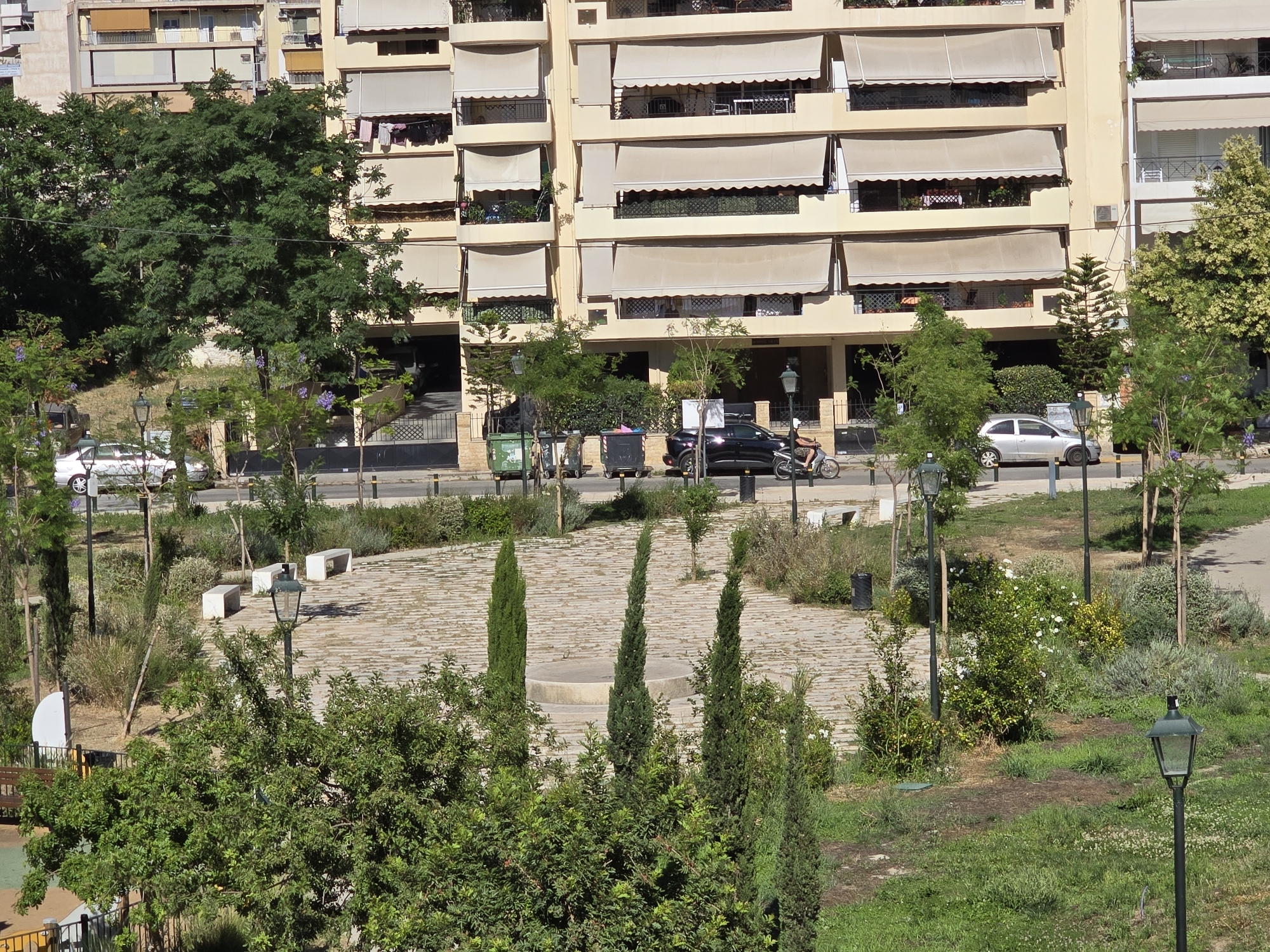 This is a high-angle exterior, daytime photograph of a street in Athens, Greece. In the foreground and midground, there is a park area with cobblestone pathways, benches, trees, and bushes. Several street lamps are visible throughout the park. In the background, there is a large, multi-story apartment building with cream-colored walls and numerous balconies covered by beige awnings. The street in front of the building has several parked cars, trash receptacles, and a person riding a scooter. The overall scene depicts a well-maintained urban environment with a blend of residential buildings and green spaces.