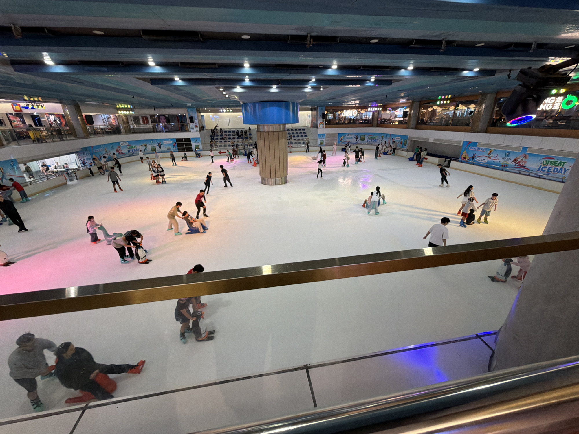 This image captures an active ice skating rink located within the Vincom Mega Mall Thao Dien in Thủ Đức, Vietnam. The scene is observed from an elevated perspective, likely from an upper floor of the mall, looking down into the rink area.
The ice rink occupies the central ground level, presenting a large, smooth white ice surface intermittently illuminated by shifting blue, pink, and purple lights originating from below or along the rink's edges. Numerous individuals, appearing to be a mix of adults and children, are engaged in ice skating. Many skaters utilize various aids: several penguin-shaped push aids are scattered across the ice, typically being pushed by younger skaters or those learning, and at least two red sled-like devices are visible, with individuals seated on them while being pushed by others.
Specific individuals and their actions include:
* In the foreground, two adults are interacting with a red sled aid; one individual is seated on it, wearing red skates, while another, wearing green skates, appears to be pushing them.
* To the left, a person in a grey shirt and shorts with teal-colored skates stands near the barrier.
* In the middle ground, several skaters are actively moving, some confidently, others cautiously. A person in a black jacket and black pants with dark skates is shown in a falling or seated position, interacting with a red sled aid.
* Another individual is pushing a black sled aid.
* Near the far barrier, individuals are seen pushing a variety of penguin-shaped aids, some standing, others in an assisting posture.
* A group of people wearing skates are congregated near the barriers, some observing, others preparing to enter or exit the ice.
The rink is enclosed by a low barrier, beyond which is a walkway and then the multi-level structure of the shopping mall. The barriers around the rink feature "UPBEAT ICE DAY" and "VINCOM" branding, confirming the location and activity. The mall environment includes multiple floors with visible storefronts and restaurants lining the perimeter above the rink, indicating a lively commercial setting. The ceiling above the rink is a series of blue-striped panels with recessed lighting fixtures. A prominent cylindrical pillar, light brown with a blue top section, stands centrally within the rink area, serving as a structural element. Overhead, metallic railings of the viewing platform are visible in the foreground, partially obscuring the lower section of the rink.