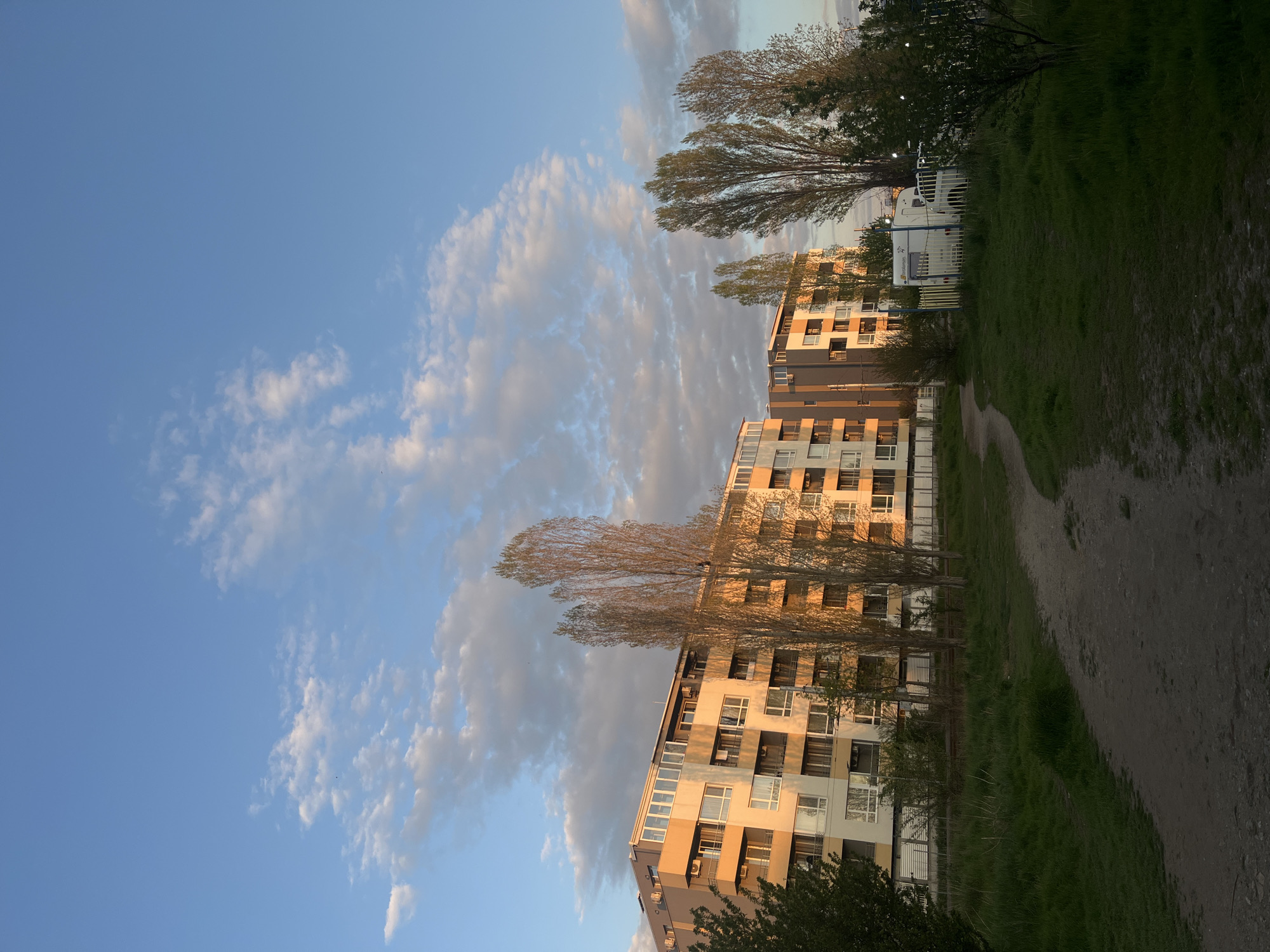 The image captures an urban-fringe landscape, likely in Sofia, Bulgaria, illuminated by the warm, low-angle light of either late afternoon or early morning (golden hour). The scene is set against a vast, clear blue sky, partially filled with scattered, wispy white clouds that reflect the golden sunlight.
On the left side of the frame, several modern multi-story residential buildings rise, their facades featuring a mix of light-colored (beige/white) and darker (grey/brown) panels. Numerous windows and balconies are visible, with some reflecting the sun's warm glow. In front of and between these buildings, tall, slender trees with relatively sparse foliage stand, suggesting either early spring or late autumn.
The right side and foreground of the image consist of a natural or semi-natural green space. A winding dirt path cuts through vibrant green grass, leading into the distance. Further back on the right, partially obscured by trees and greenery, a white caravan or mobile home-like structure on a trailer is visible, enclosed by a white fence. The overall impression is peaceful, with the warm light highlighting the architectural and natural elements.
No specific human activity is depicted. Visible text on the white caravan or fence is present but not clearly legible.