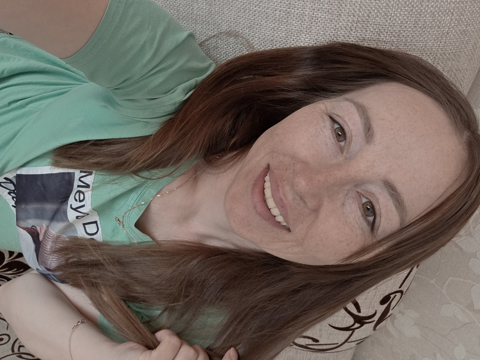 A female individual is depicted in a close-up selfie, smiling directly at the camera. She has long, brown hair, fair skin with visible freckles across her nose and cheeks, and brown eyes. Her upper teeth are visible as she smiles. She wears a light green, short-sleeved t-shirt featuring a partially visible graphic print with text and an image of a person. Around her neck is a thin gold-colored necklace with a small pendant, and a delicate gold-colored bracelet adorns her left wrist. Her left hand is observed gently grasping a strand of her own hair near her left shoulder. The background indicates an indoor residential setting, with a light beige woven fabric surface visible on the left and a light-colored fabric with a dark, ornamental pattern on the right, consistent with upholstered furniture. This scene is located in the city of Chaikovsky, Russia.