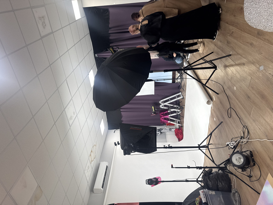 An indoor photography studio is depicted in Sofia, Bulgaria, featuring a light wood laminate floor and a drop-tile ceiling with multiple water stains. A white seamless paper backdrop is rolled out from the left wall, extending onto the floor. Several pieces of photographic lighting equipment are positioned throughout the room, including a large black studio umbrella on a stand in the right foreground, a black softbox on a stand near the center, and a smaller pink-domed light on a stand to the left. A floor-standing spotlight is visible on the left foreground. Electrical cables are sprawled across the floor, connecting various devices.
Four individuals are present. In the mid-ground, a person with light pink hair, wearing a black long dress with an open back and black sneakers, stands facing away from the camera. To their right, another person in a long tan coat is partially visible, also facing away. Further left and slightly behind the pink-haired individual, a person in dark clothing and light-colored shoes is crouching near the white backdrop. A fourth person, also in dark clothing, is partially visible in the background interacting with an unseen element behind the white backdrop.
In the background, purple and black drapes serve as an additional backdrop. Two step ladders, one pink and one silver, are positioned against the purple drapes. An air conditioning unit is mounted high on the left wall, beneath a cluster of water stains on the ceiling tiles. A black trash bag and a clear storage bin are located in the bottom left corner, while a red bag lies near the ladders.